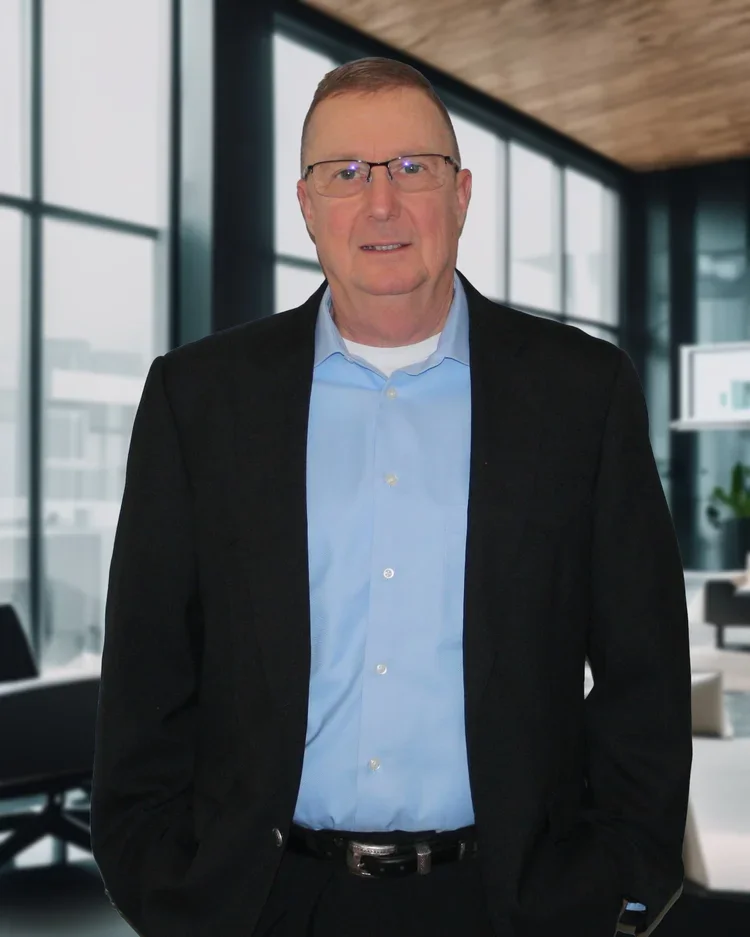 A middle-aged man wearing glasses, a light blue dress shirt, and a black blazer, standing in a modern office with large windows.