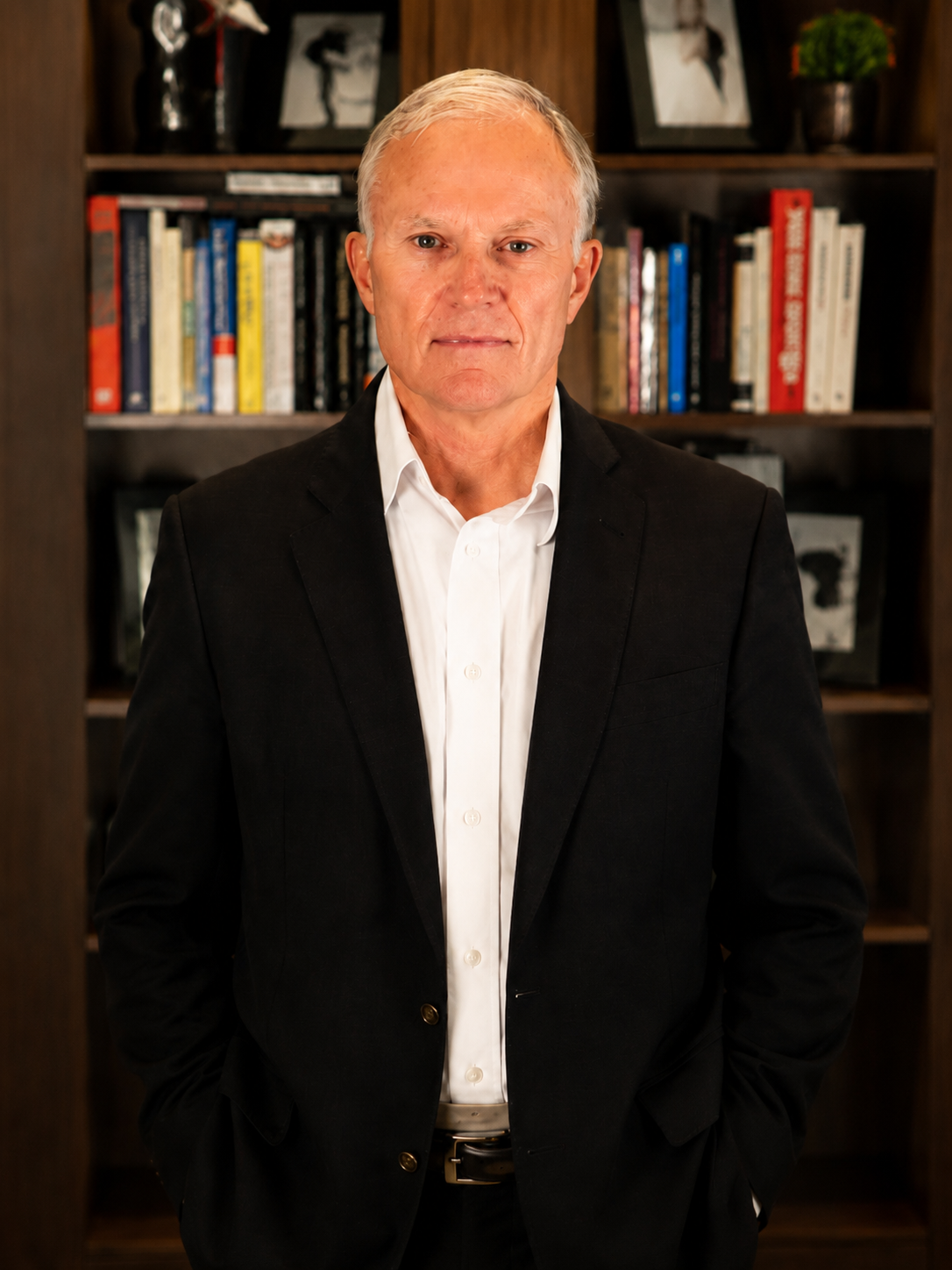 A middle-aged man with short, gray hair and blue eyes wearing a white dress shirt and black blazer, standing in front of a bookshelf filled with books and framed photos.