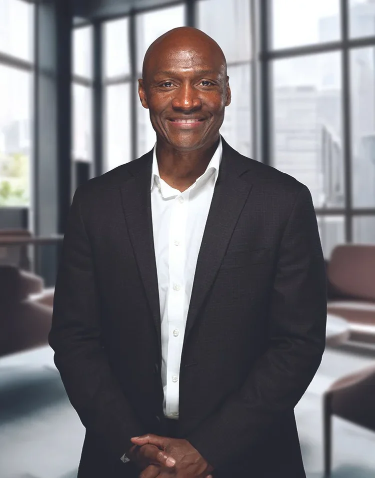 A smiling man wearing a dark suit and white shirt, standing in a modern office with large windows and urban cityscape background.