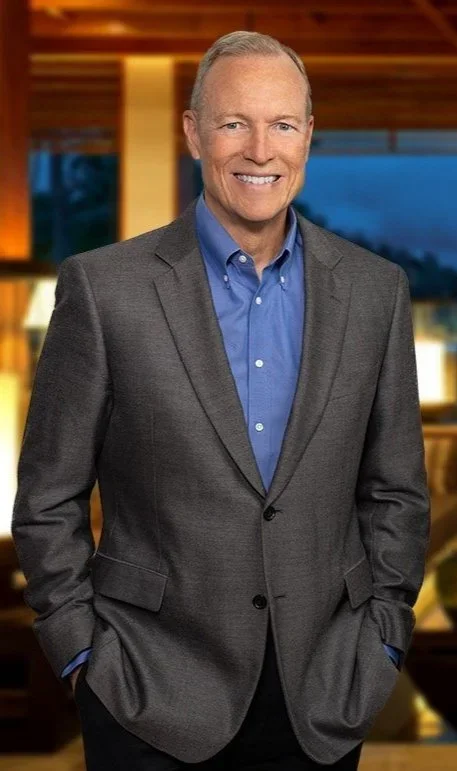 A smiling middle-aged man in a gray suit and blue dress shirt, standing indoors with warm lighting and wooden decor in the background.