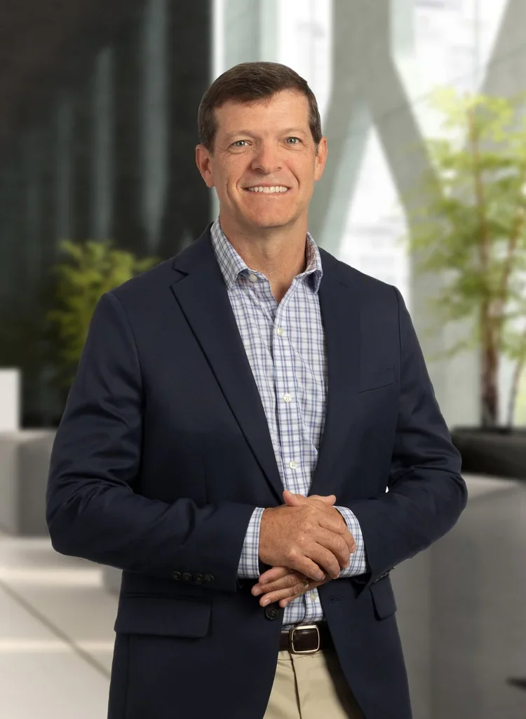 A man in a dark blazer and checked shirt standing indoors with modern architecture and green plants in the background.