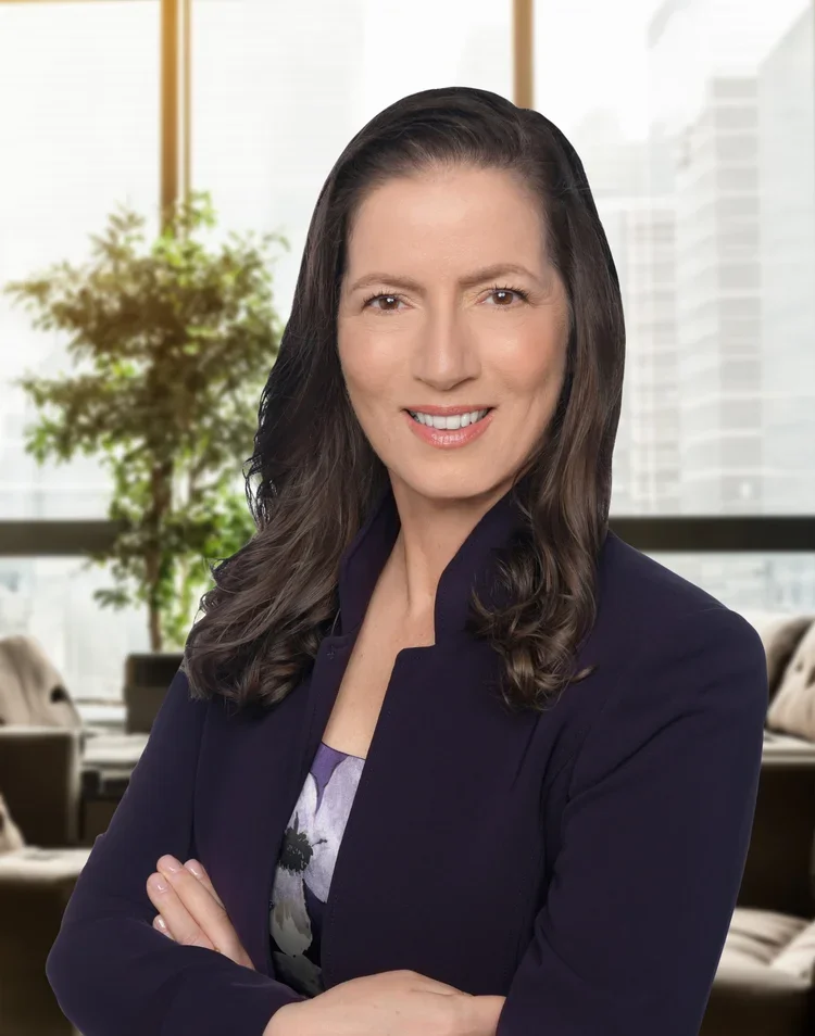 A professional woman with long dark hair, wearing a dark blazer and patterned top, standing with arms crossed in a modern office with large windows and a potted plant in the background.