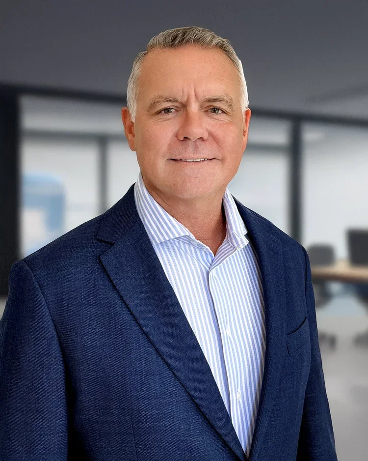 A middle-aged man with short, gray hair wearing a dark blue suit jacket and a light blue and white striped dress shirt, standing in a blurred modern office setting.