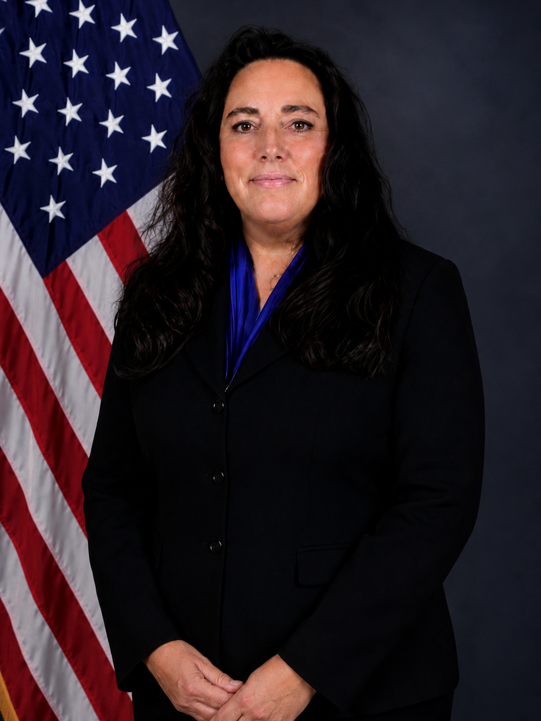 A woman with long dark hair, wearing a dark blazer and blue blouse, standing in front of an American flag.