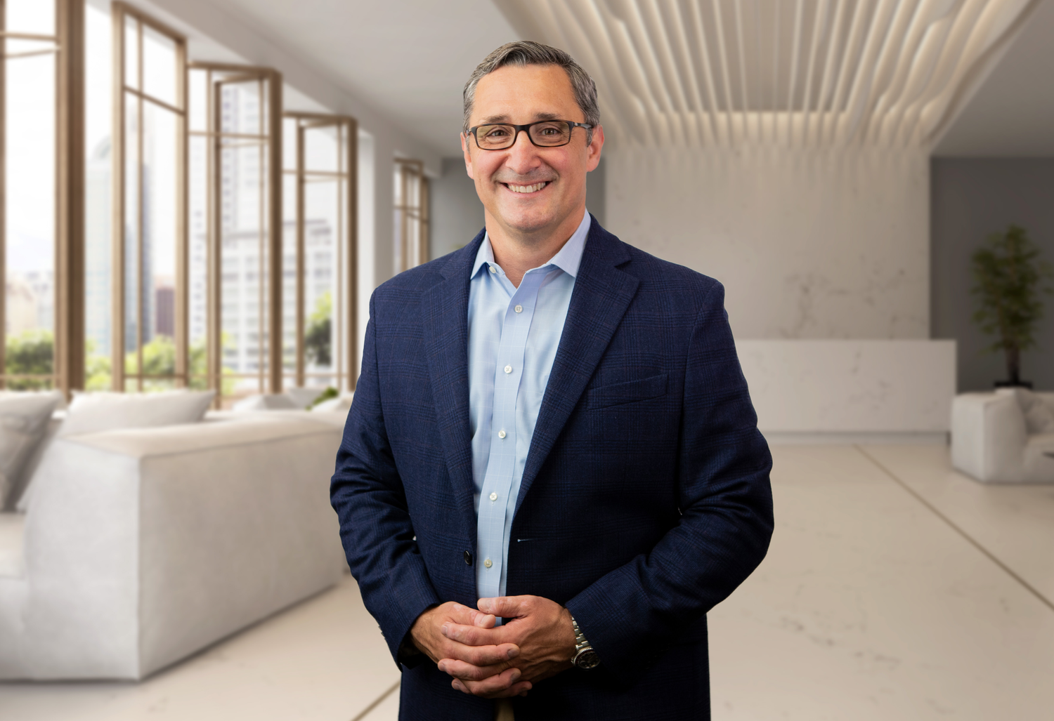 A man in a blue suit and glasses standing in a modern, bright office or lobby with large windows and city views.