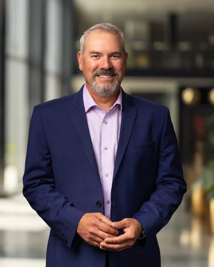 A middle-aged man with gray hair and a beard wearing a navy blue suit and light purple dress shirt, standing indoors with a blurred background.