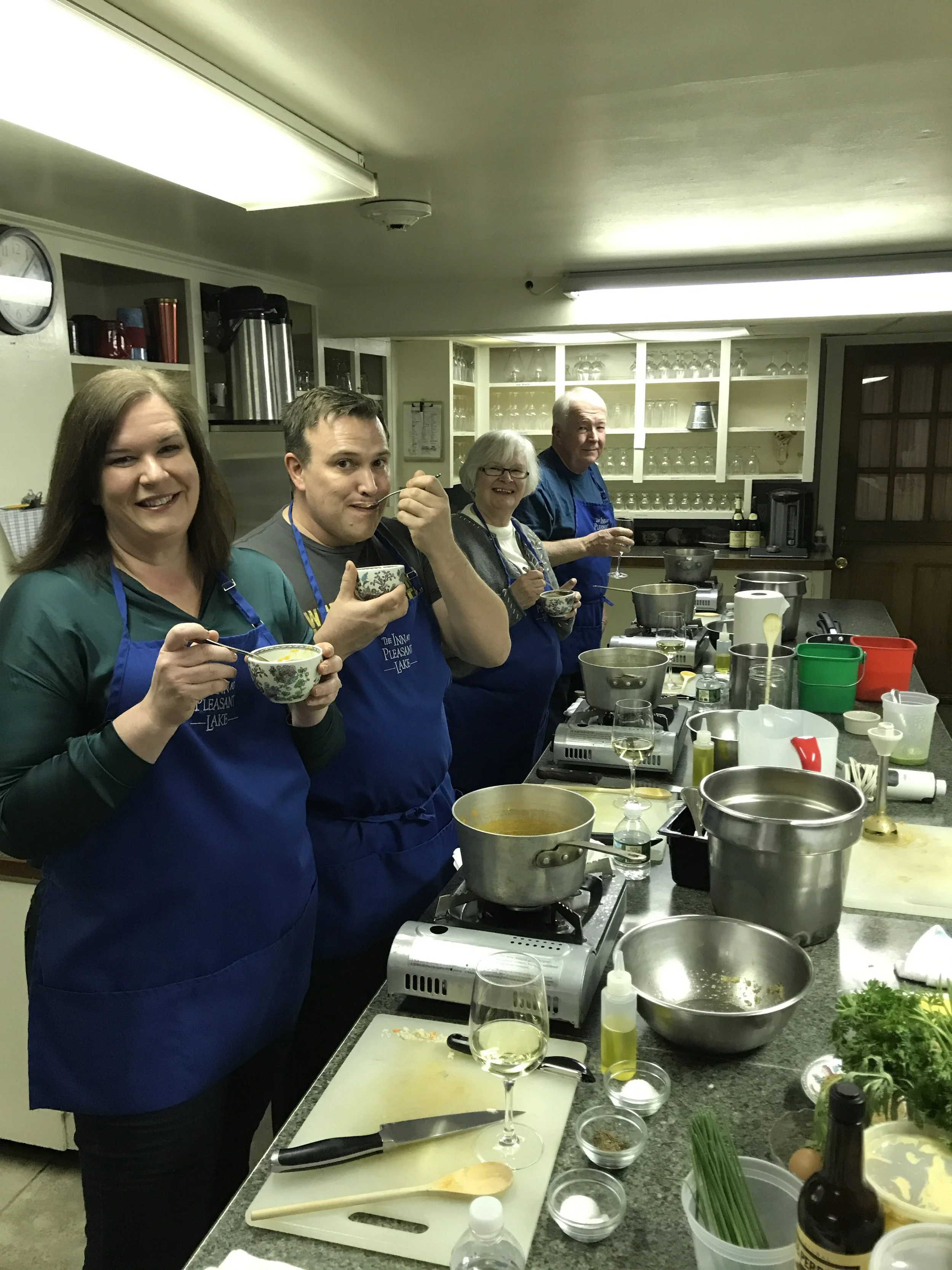 A group of four in blue aprons tasting from bowls at the kitchen counter.