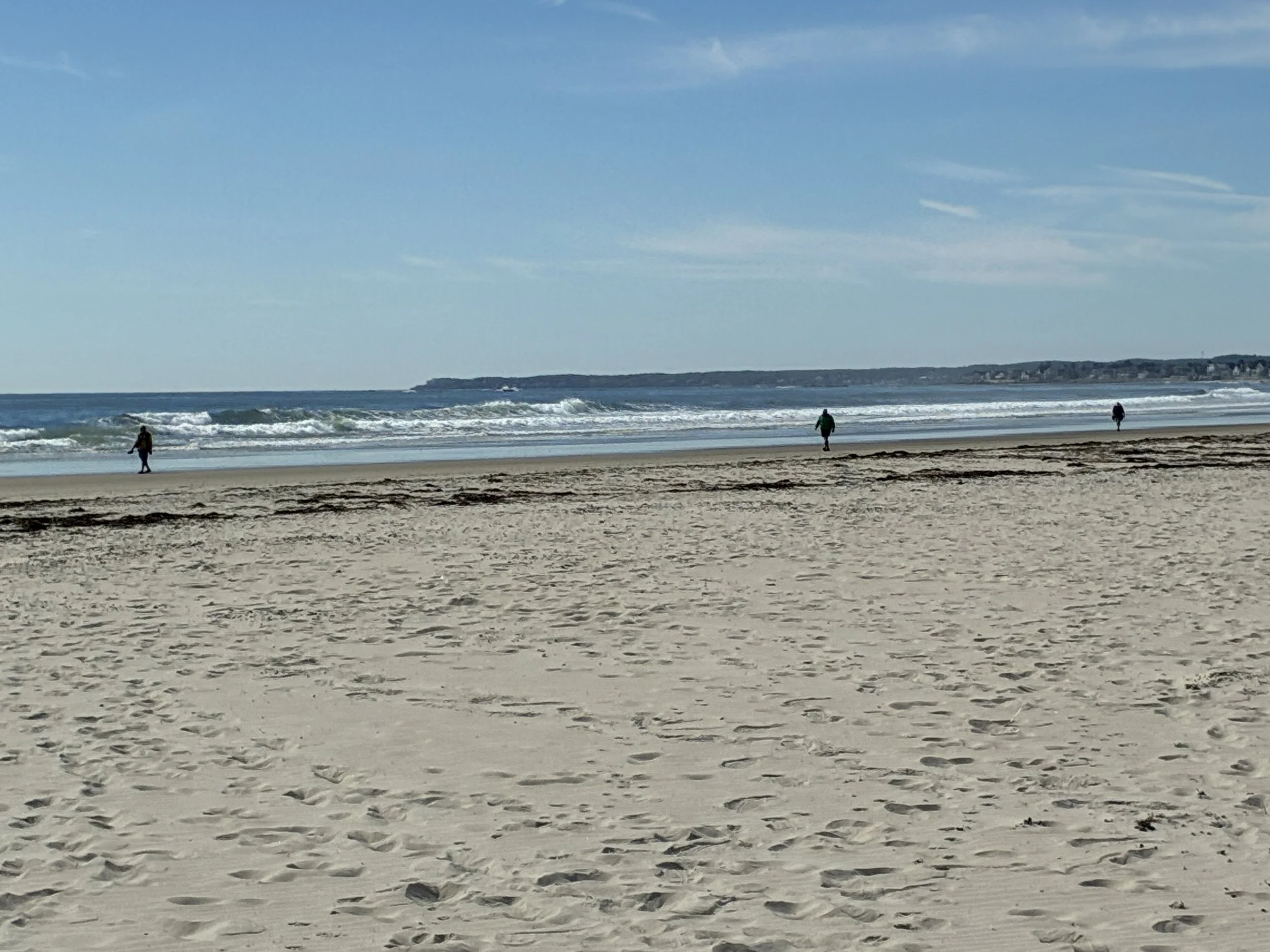 A wide sandy beach in fall with people walking along the surf.