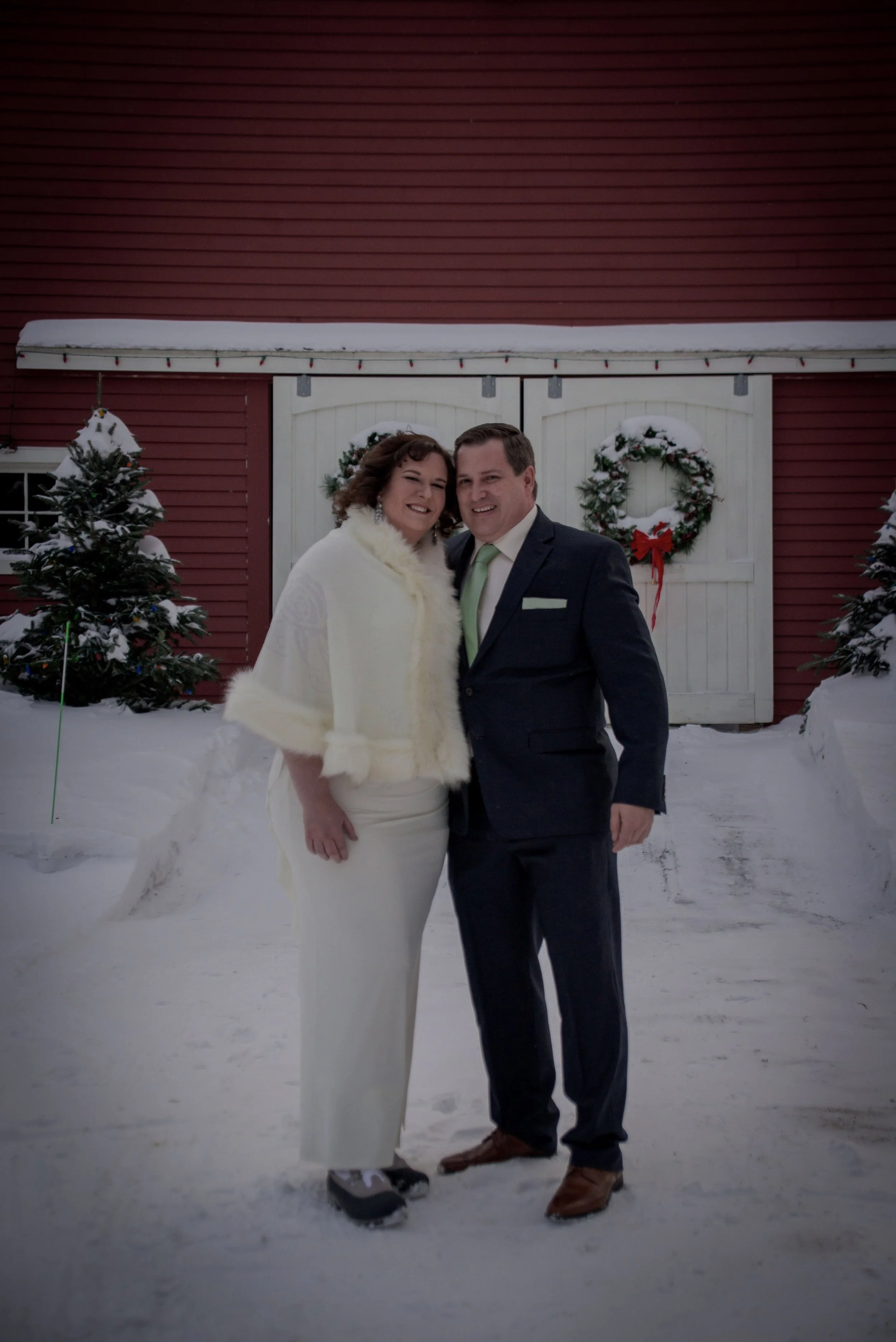 A bride and groom in the snow in front of the inn's red barn with wreath-decorated doors.