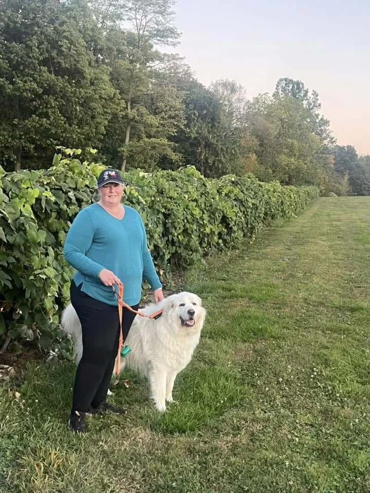 A woman with a leashed white dog beside a grapevine row.