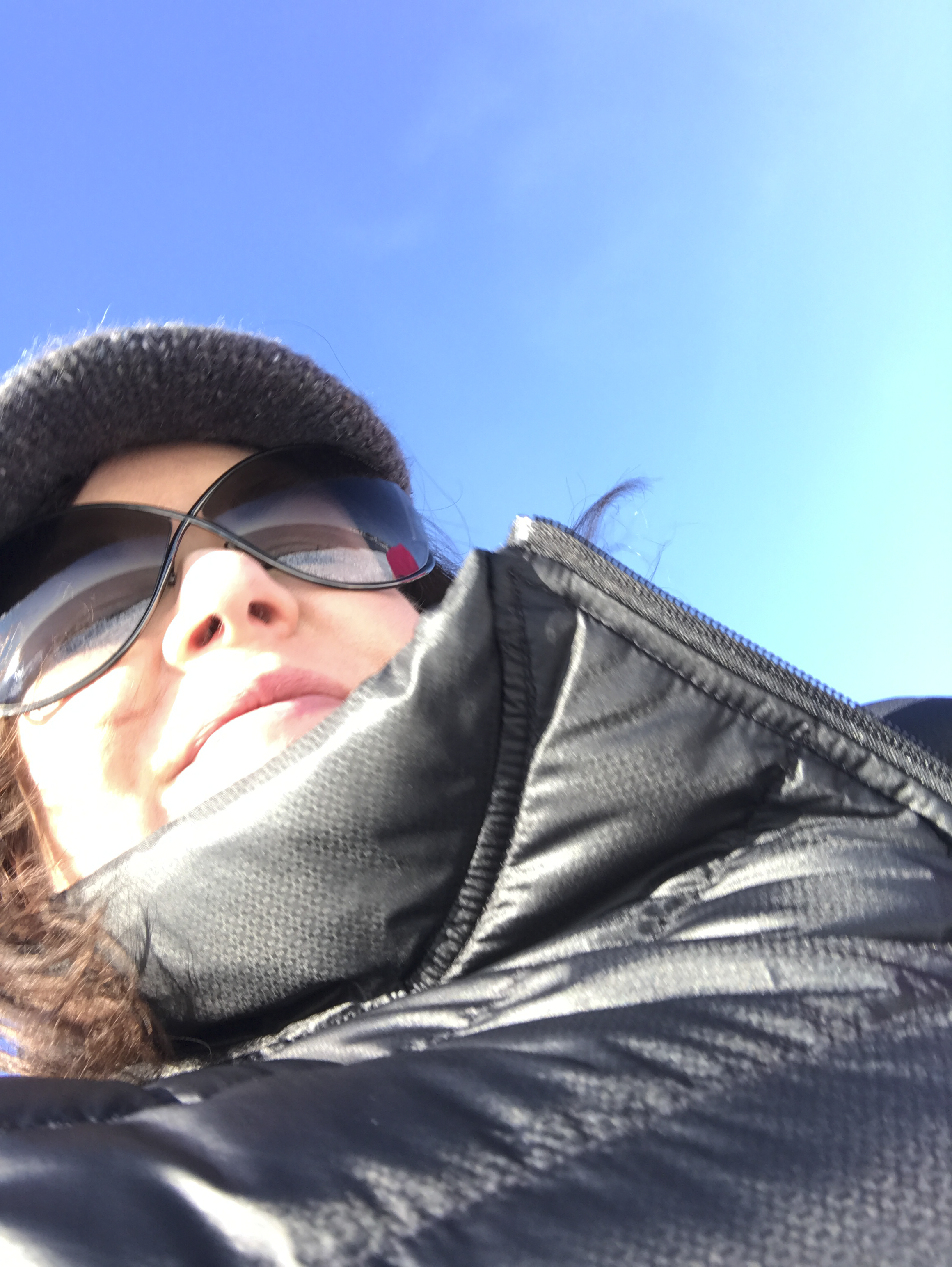 Close-up of a person wearing sunglasses, a black beanie, and a black jacket, with a clear blue sky in the background.