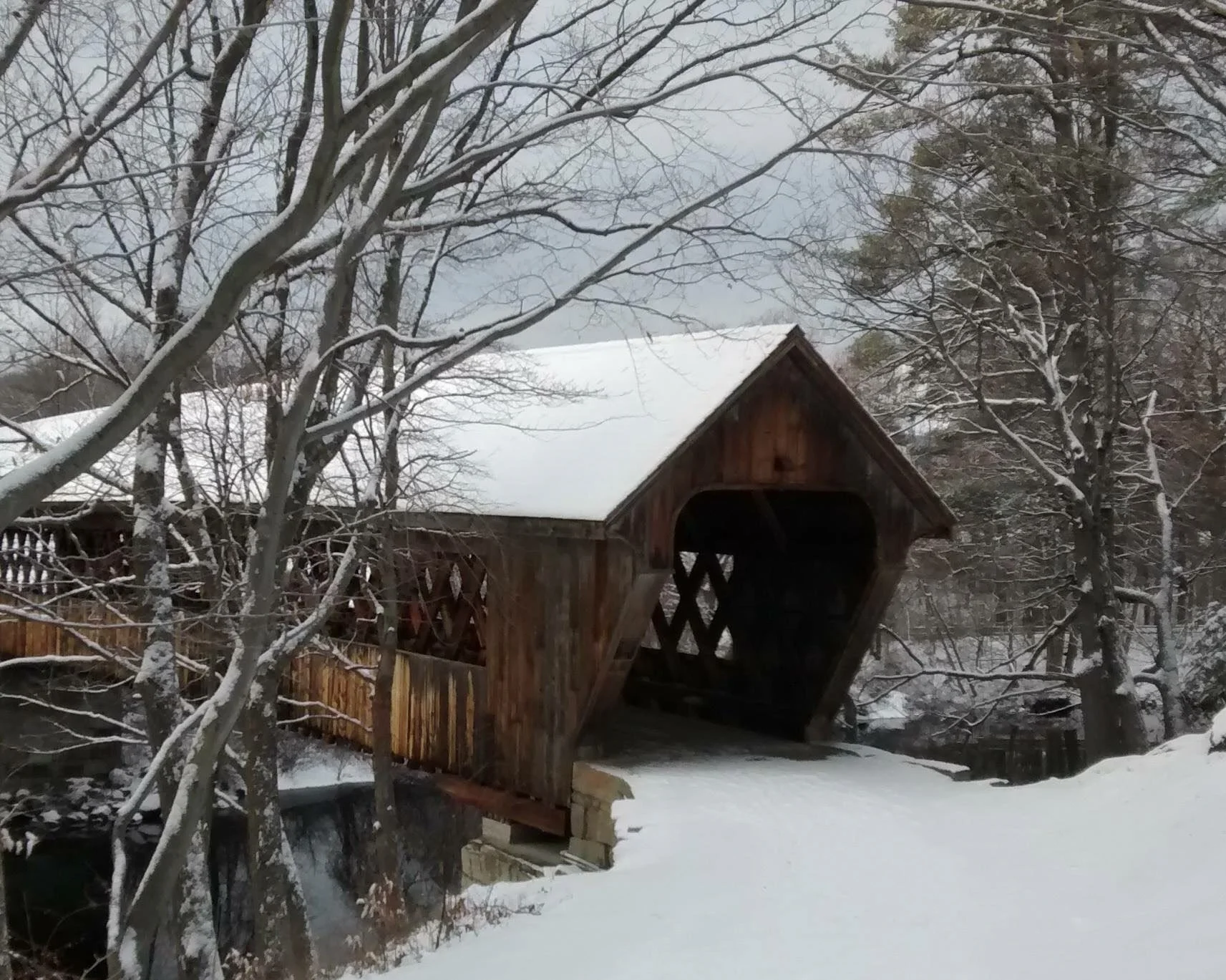 Frozen winter scene with a snow-covered wooden covered bridge surrounded by trees with snow-laden branches.