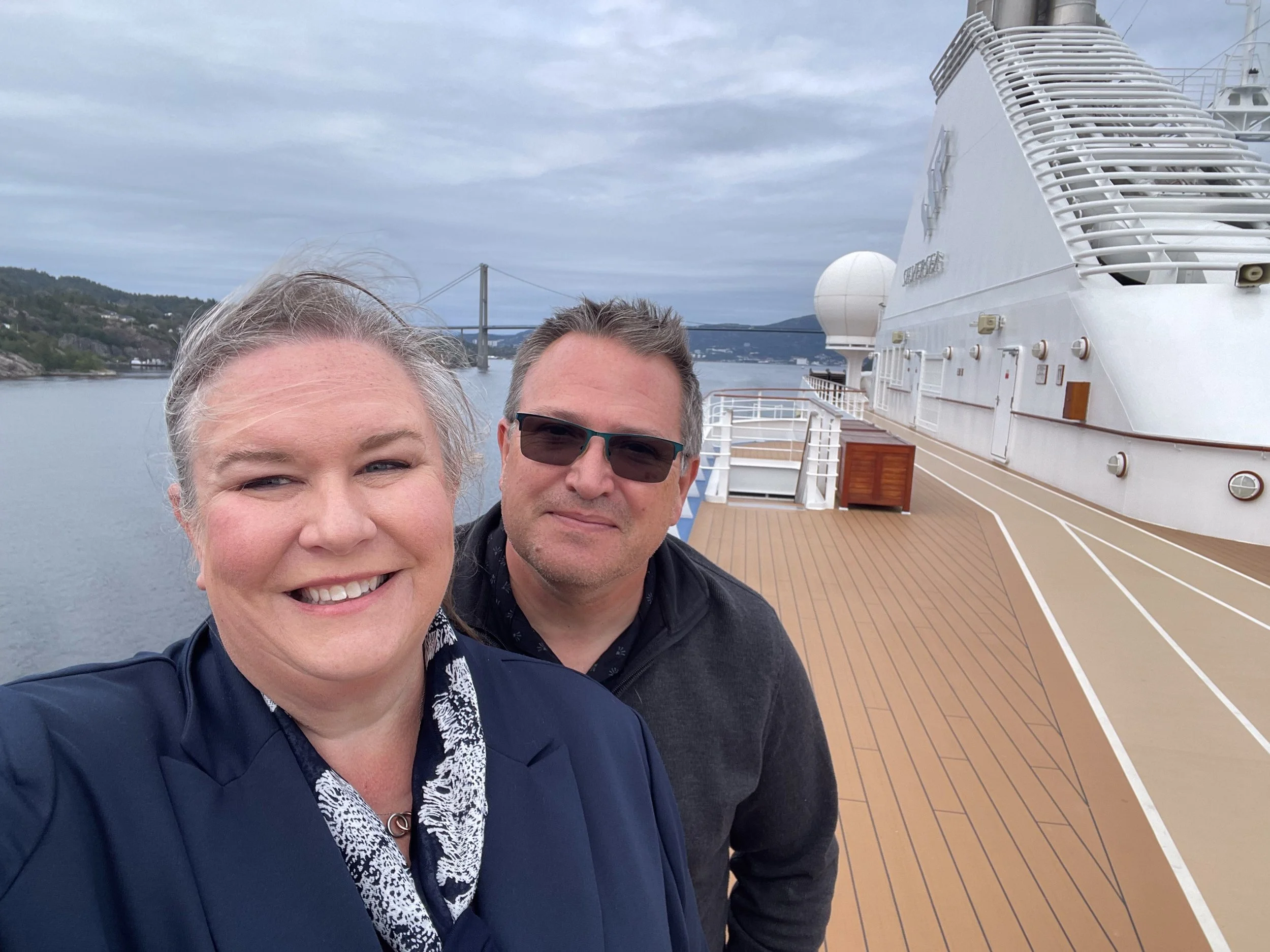 Smiling man and woman taking a selfie on a cruise ship deck with a large white ship and a bridge over water in the background.