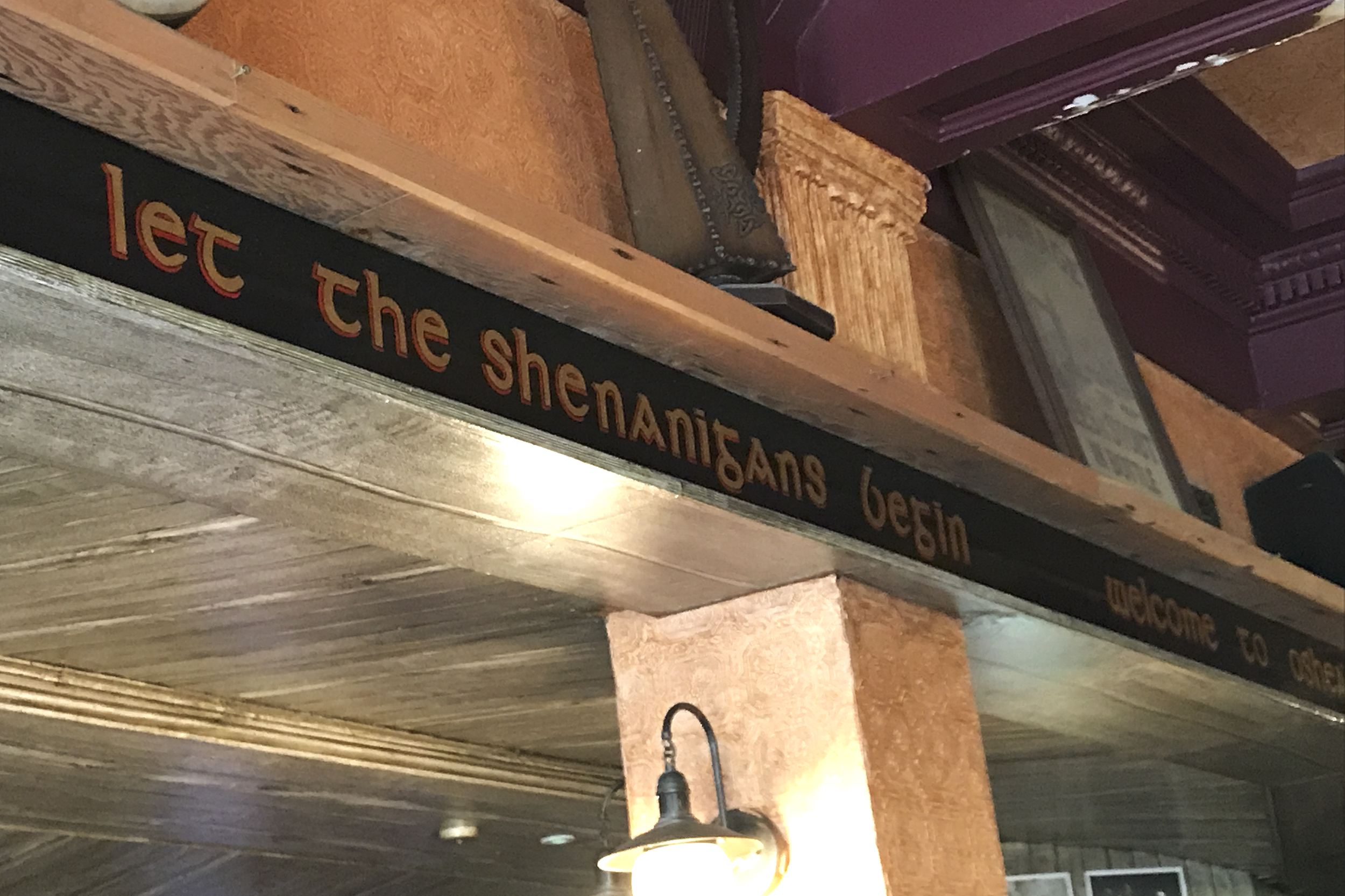 Close-up of a black metal sign with orange lettering reading 'Let the shenanigans begin.' partially visible, mounted on a wooden wall, with a ceiling light and decorative molding in the background.