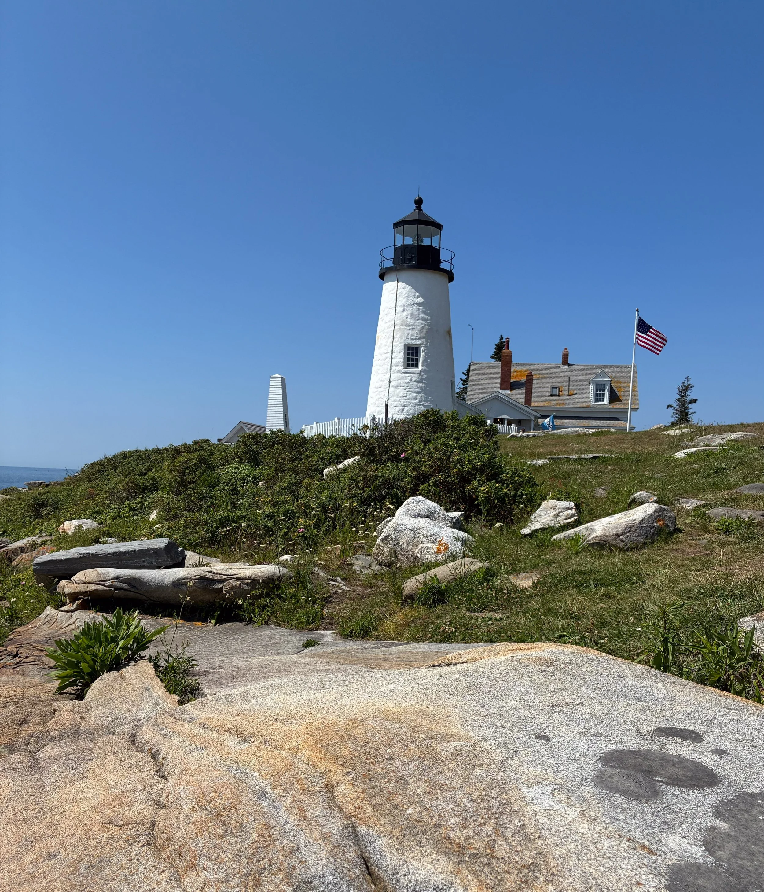 A white lighthouse with a black top stands on a grassy hill near the coast, with a blue sky and an American flag flying nearby.