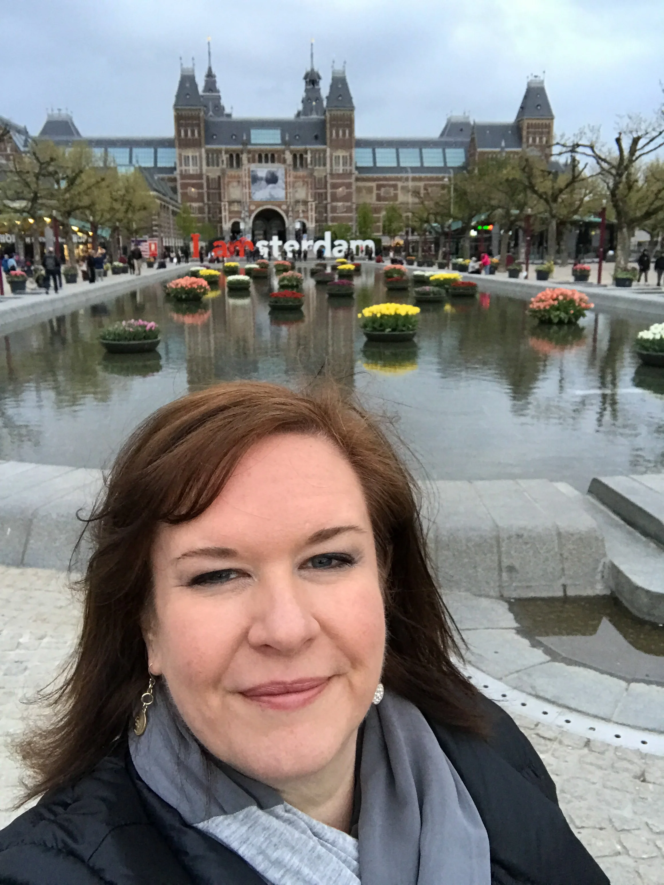A woman taking a selfie in front of the National Rijksmuseum in Amsterdam, with the 'I amsterdam' sign and flowers reflection in a pond.