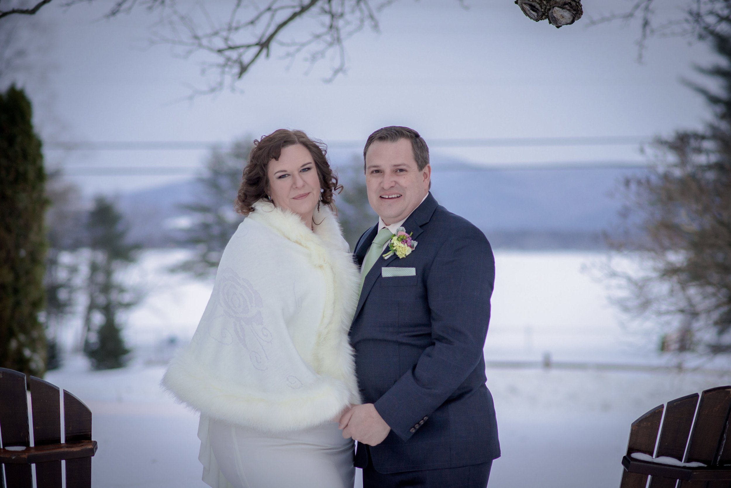 A bride and groom portrait with frozen Pleasant Lake and Adirondack chairs behind them.