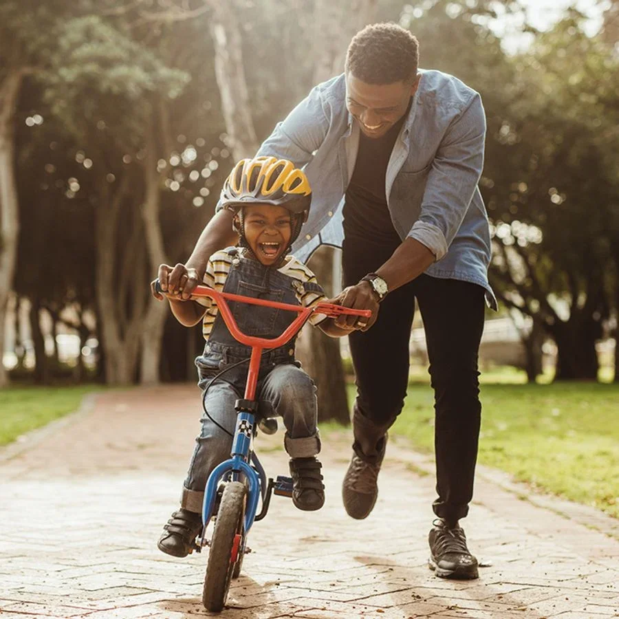 A young boy wearing a yellow helmet riding a blue and orange bicycle, laughing as an adult man helps him in a park with trees in the background.