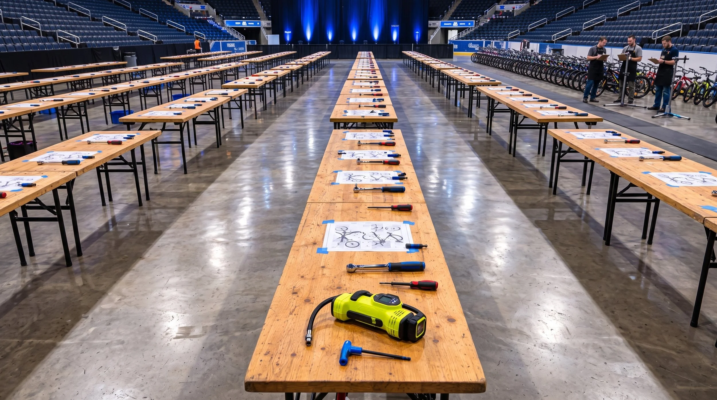 An indoor arena with rows of tables set up for a bicycle assembly or repair event, with tools and bike diagrams on the tables, and a group of people working on bicycles in the background.