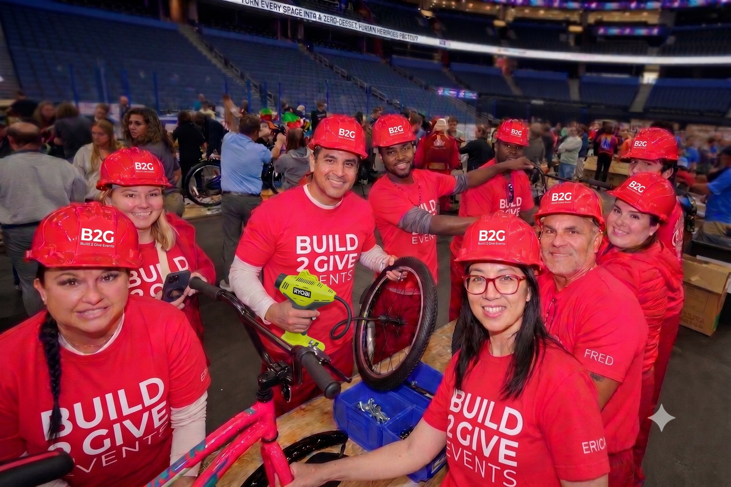 Group of volunteers wearing red shirts and hard hats, gathered inside a gymnasium, preparing for a charity event, with some holding bikes and tools.