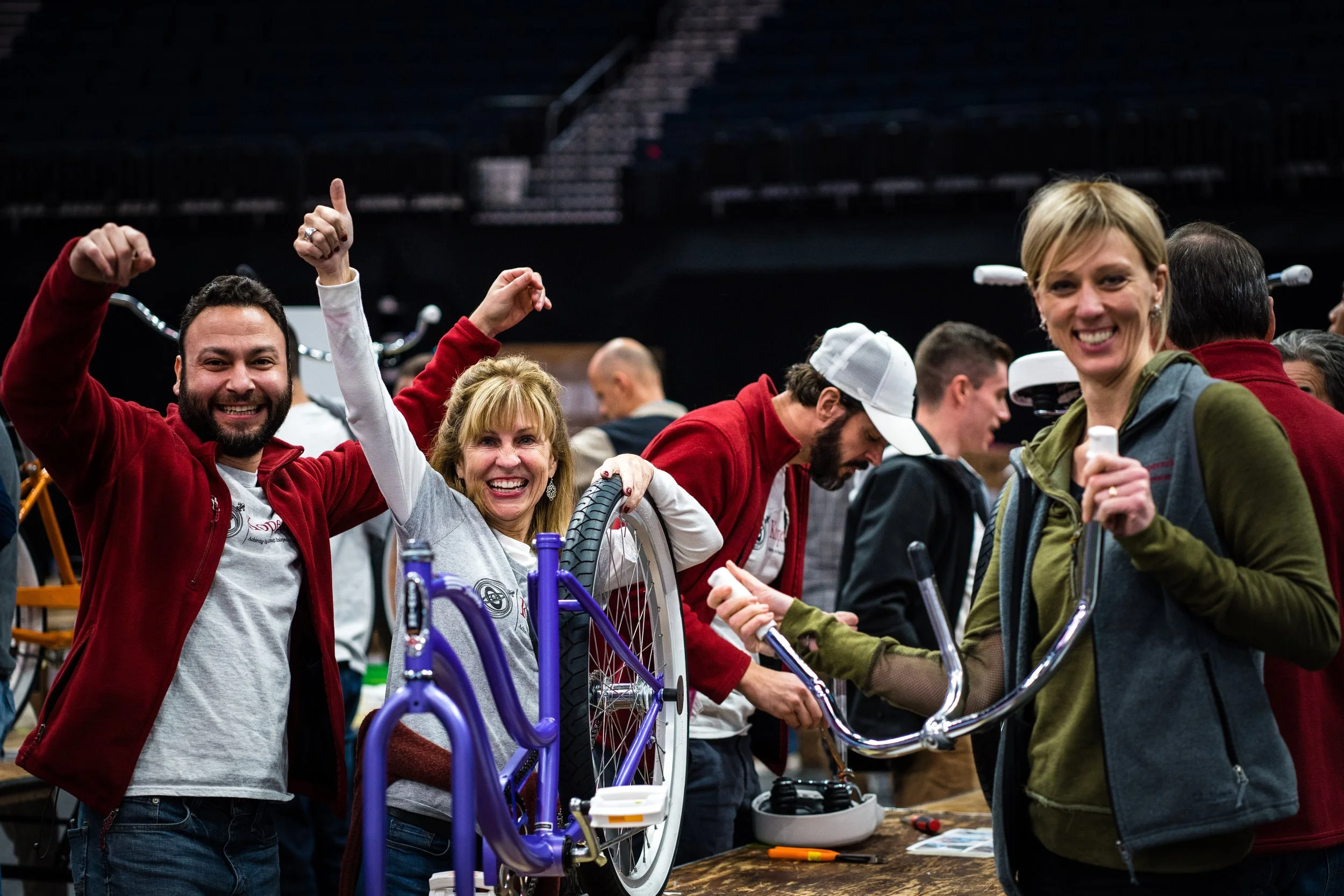Group of people at an indoor event assembling bicycles, smiling and interacting, with a woman in a green jacket holding a bicycle handlebar and others wearing red jackets