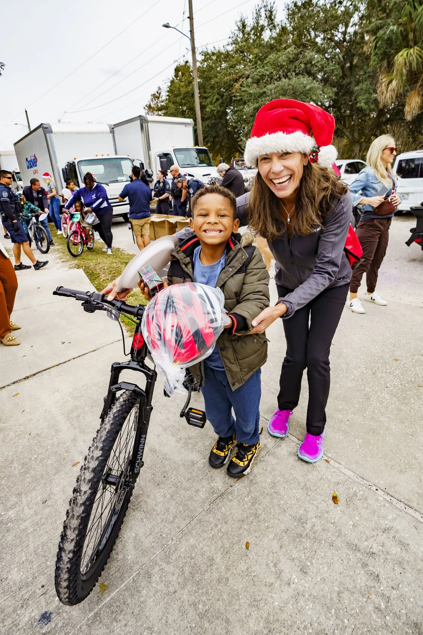 A woman wearing a Santa hat and pink sneakers smiling next to a boy on a bicycle, at an outdoor event with trucks and people in the background.