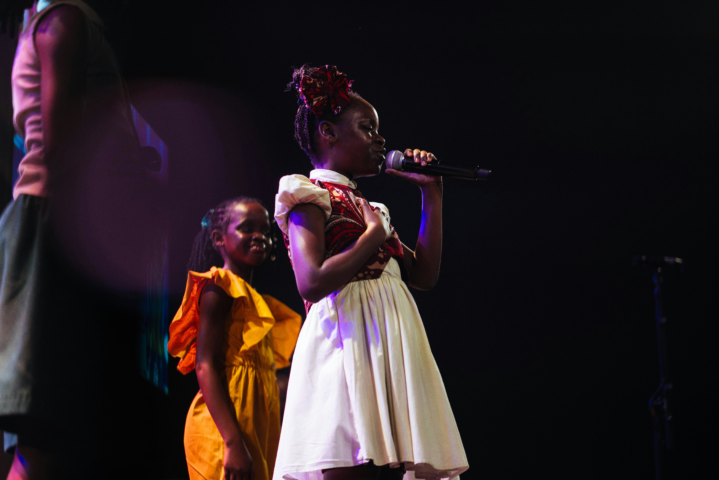 A young girl in traditional attire singing into a microphone on stage, with other children in colorful clothing standing behind her, under dim stage lighting.