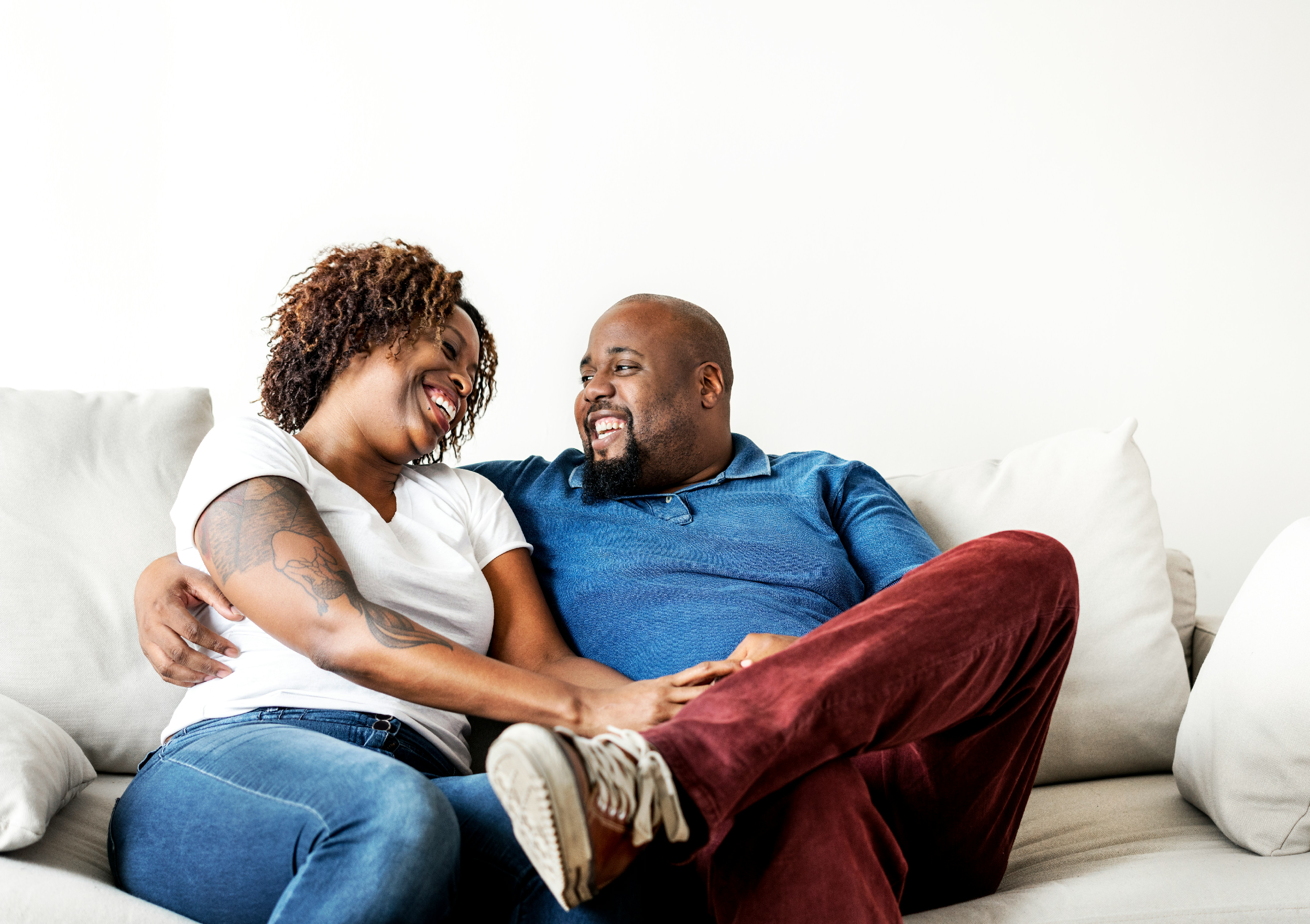 A smiling couple sitting on a white sofa, looking at each other affectionately, with one woman in a white shirt and the man in a blue shirt, against a plain white wall.