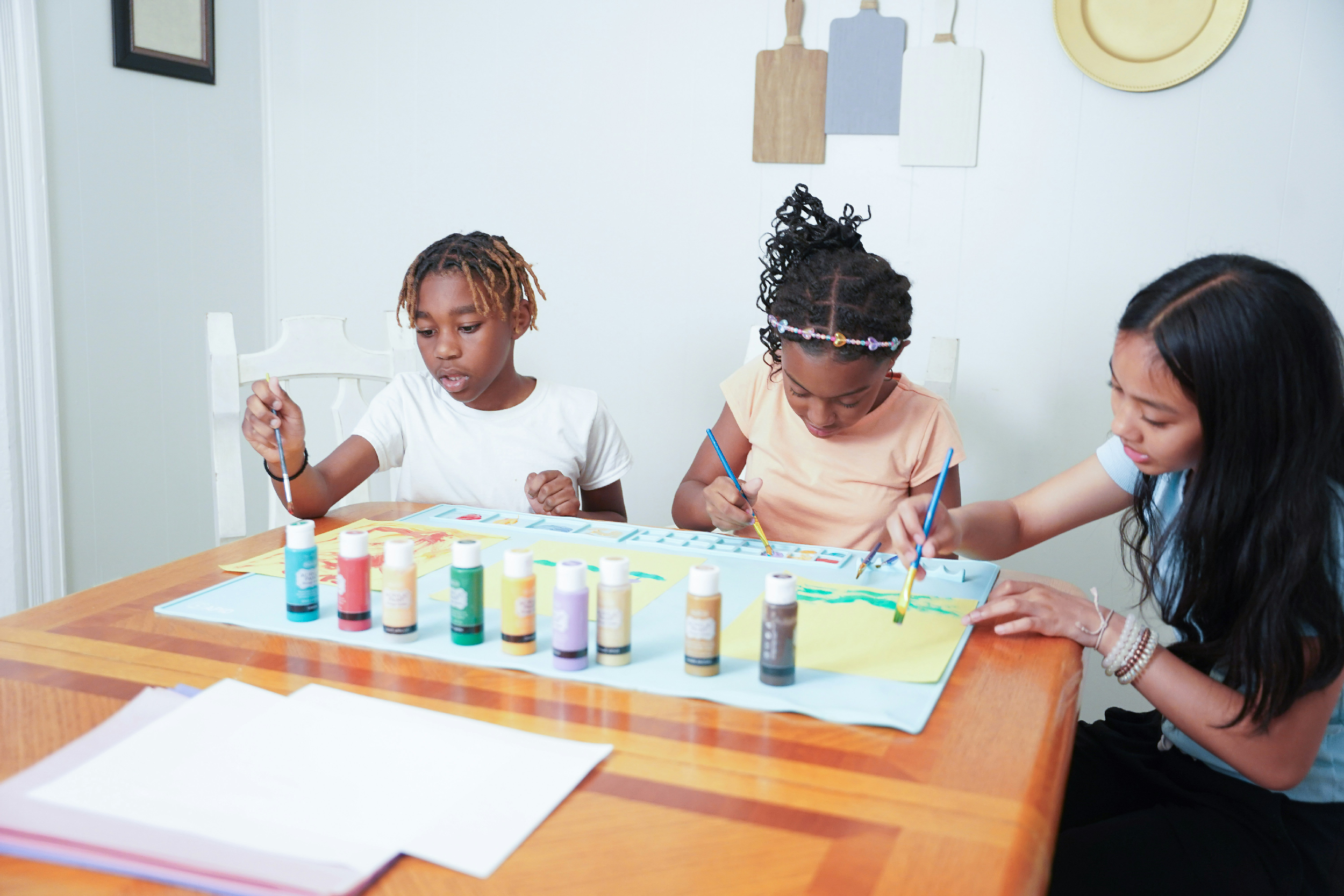 Three children sitting at a wooden table engaged in a painting activity with watercolor paints, brushes, and paper, in a decorated room with wall paintings and kitchen utensils hanging on the wall.