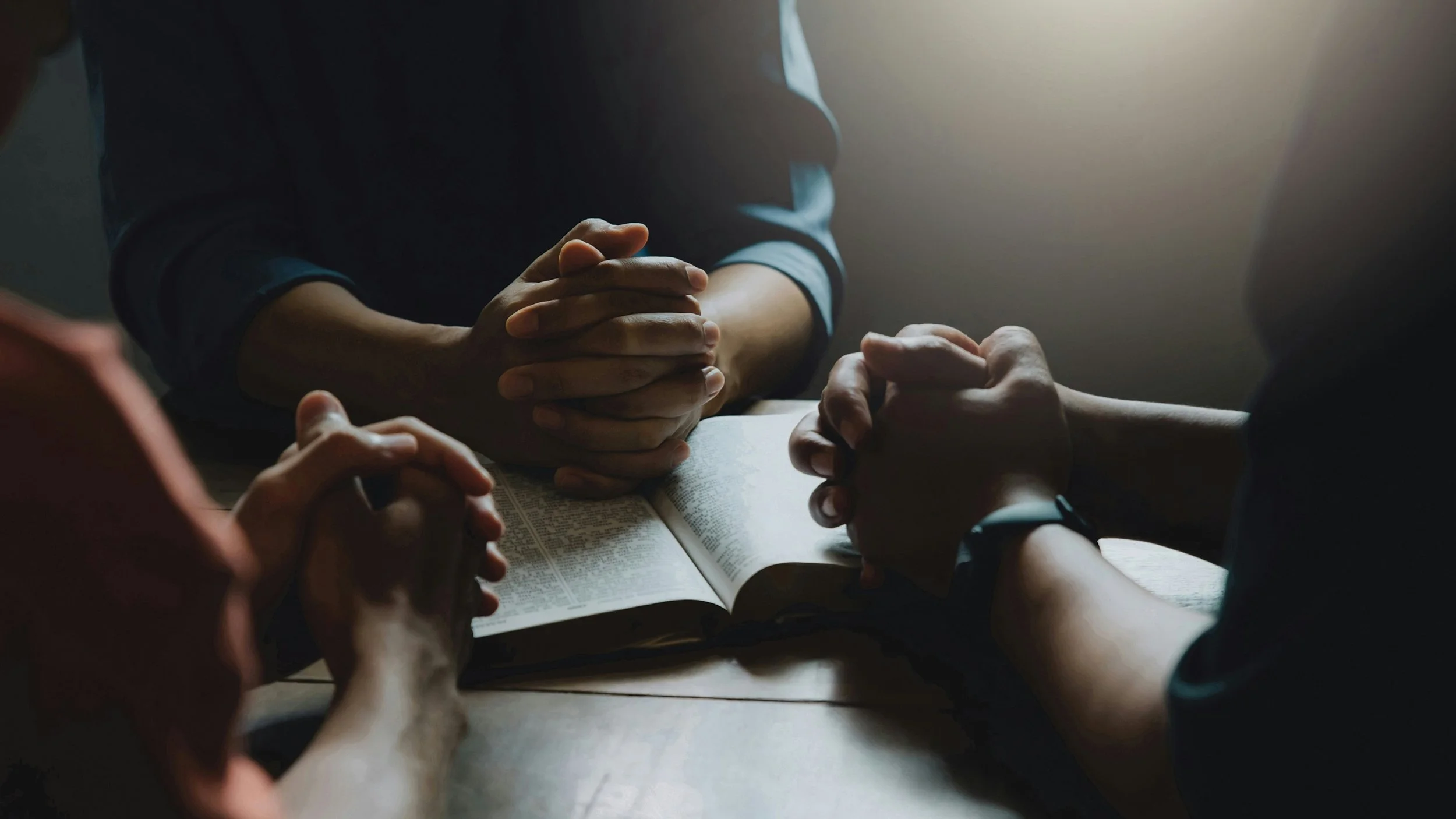 People sitting around a table with hands clasped, a Bible open in the center, engaging in a prayer or discussion.