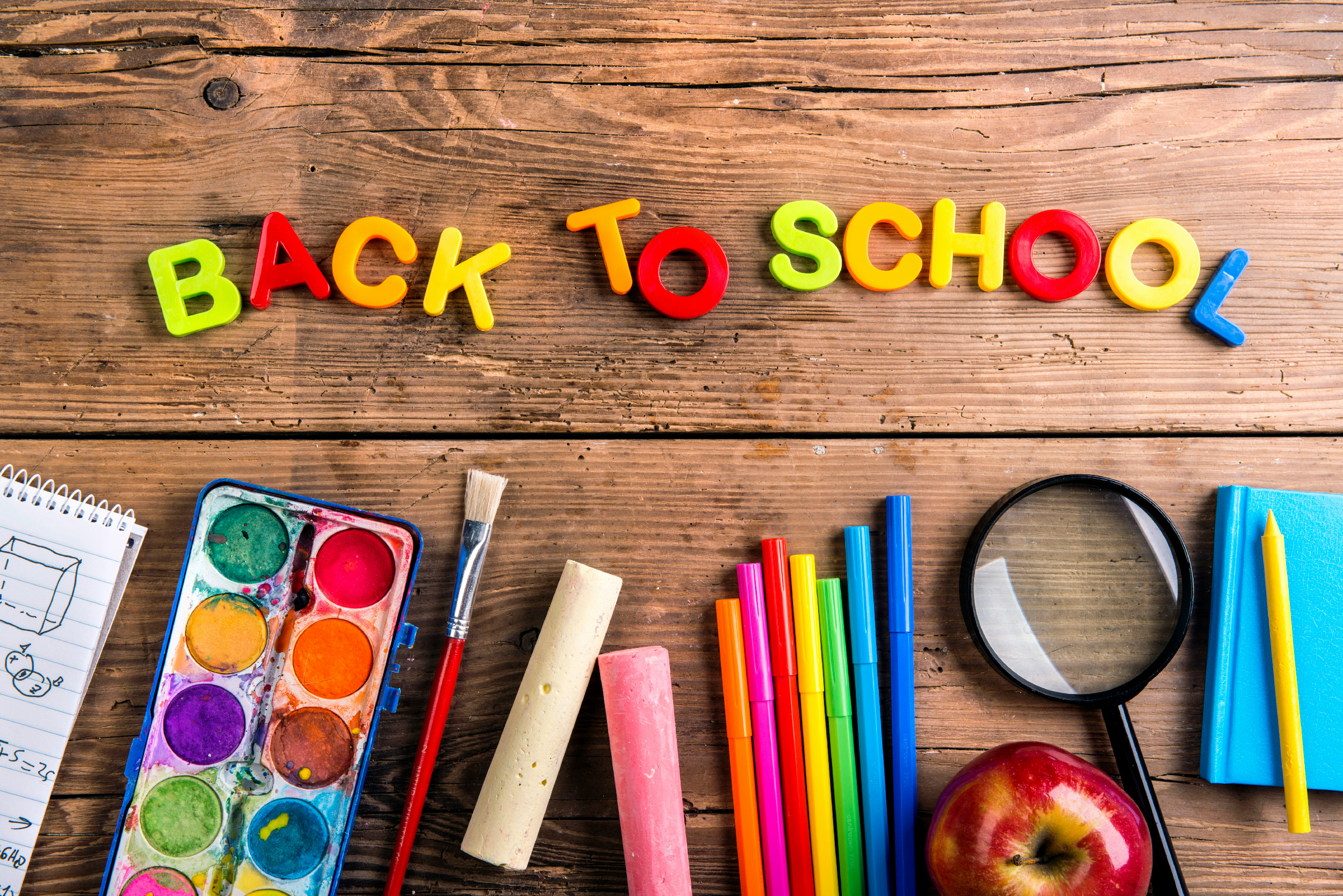 Colorful art supplies arranged on a wooden surface, including watercolor paints, markers, and a magnifying glass, with the phrase 'Back To School' spelled out with colorful plastic letters.