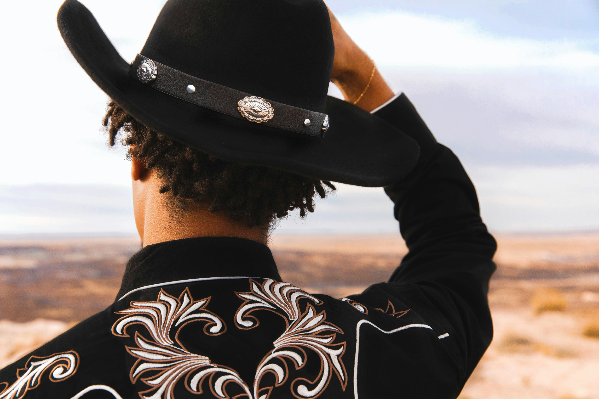 Person with short curly hair wearing a black wide-brimmed hat with decorative silver conchos and a black shirt with embroidered white and brown scrollwork, standing outdoors during sunset or sunrise with a blurred landscape in the background.