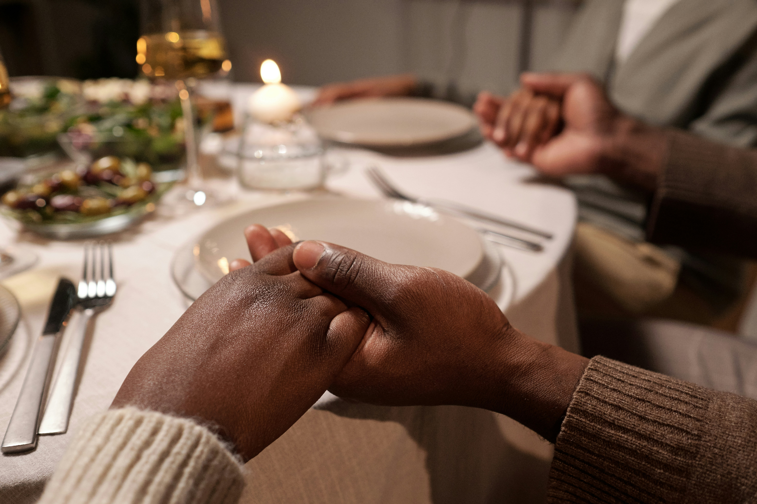 A dinner table setting with two people holding hands, with a candle, salad, and plates on the table.