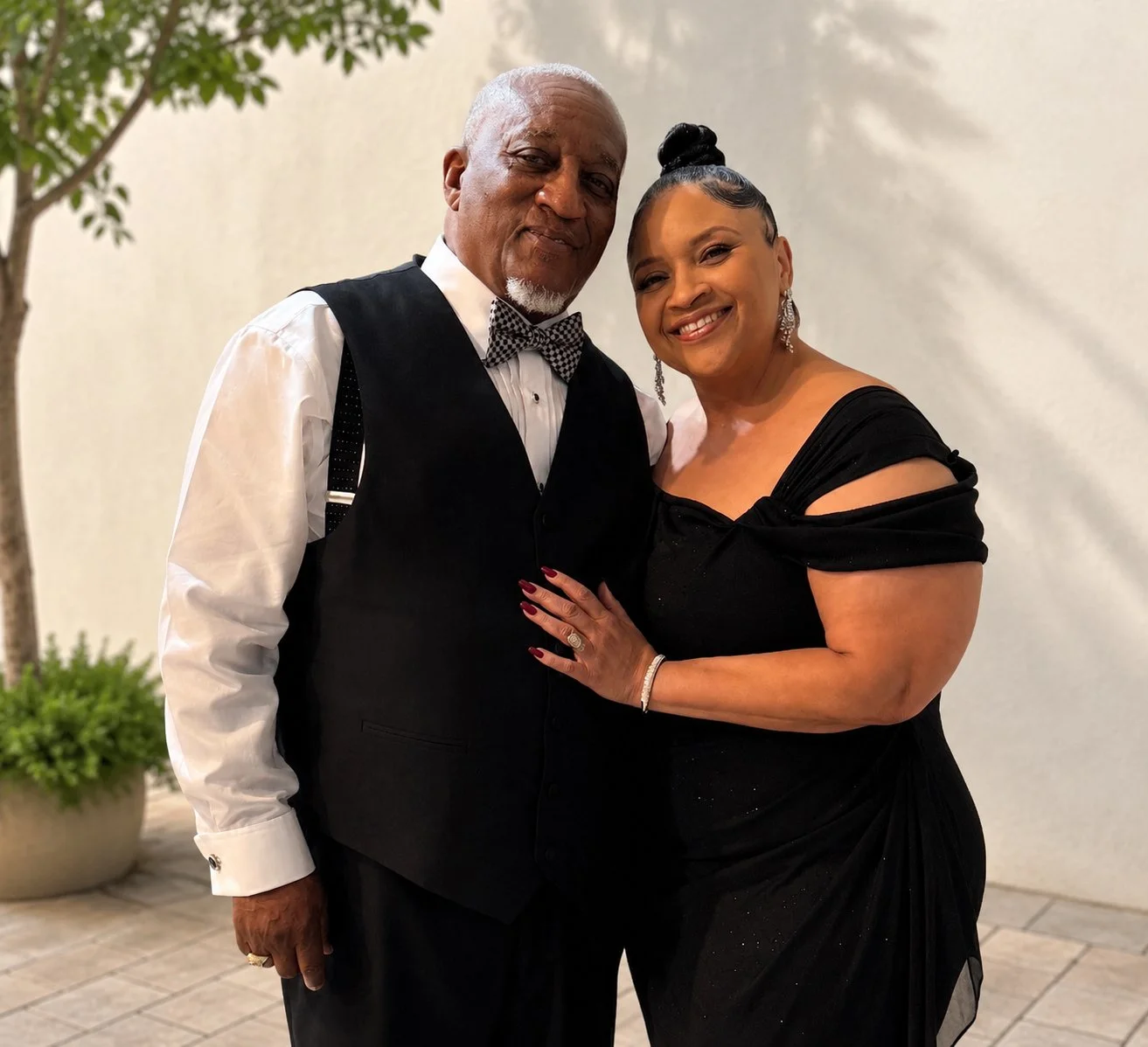 A man and a woman dressed in formal evening wear, smiling and posing together indoors. The man is wearing a black vest, white shirt, and patterned bow tie. The woman is wearing a black off-the-shoulder dress with jewelry.