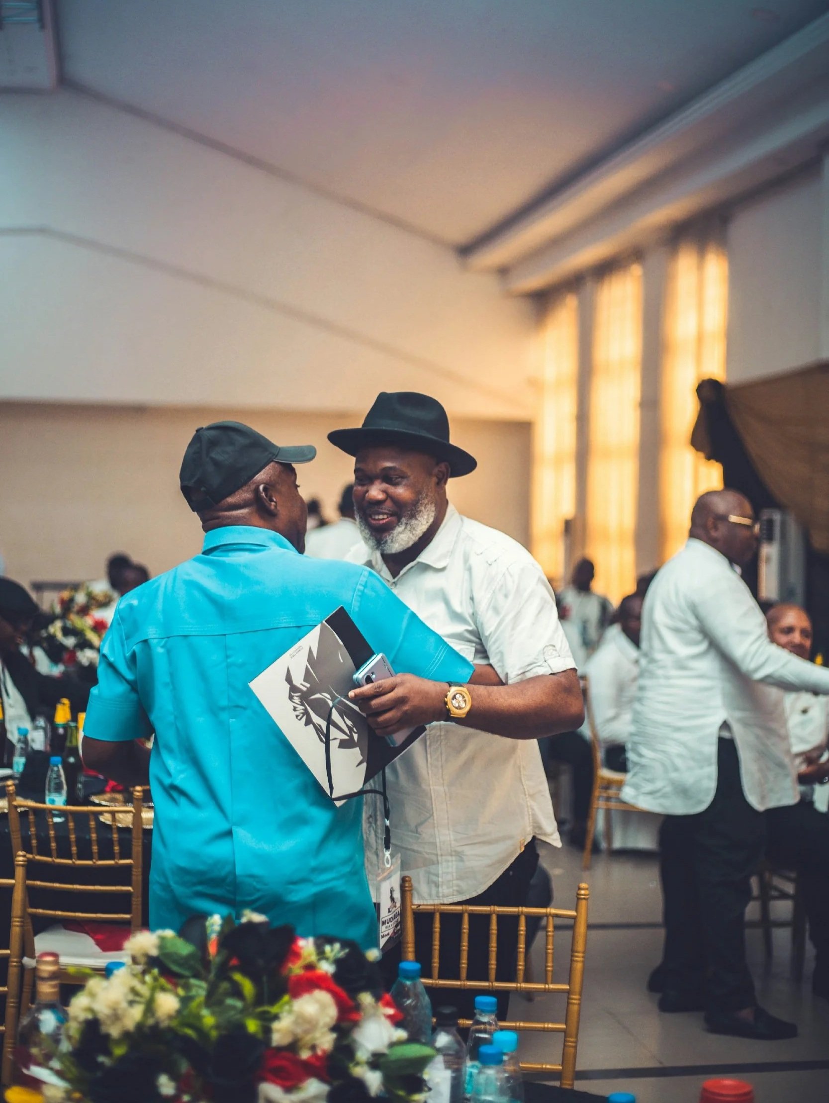 Two men are smiling and talking at a social gathering or event, with one holding a booklet and a phone, and surrounded by other attendees in a well-lit indoor setting with large windows and decorated tables.