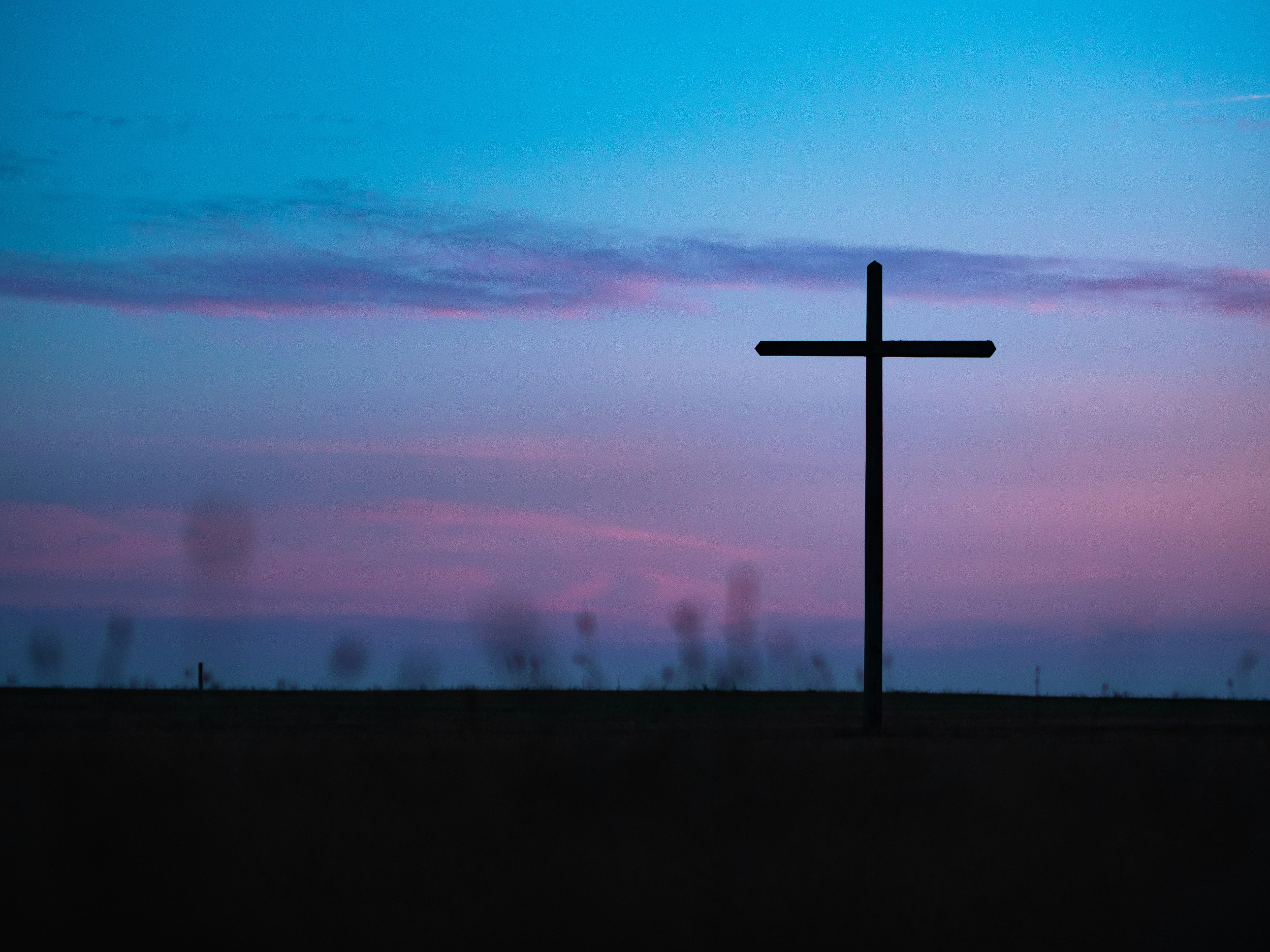 A black cross standing in a field during sunset or twilight, with a sky filled with scattered clouds in shades of blue, purple, and pink.