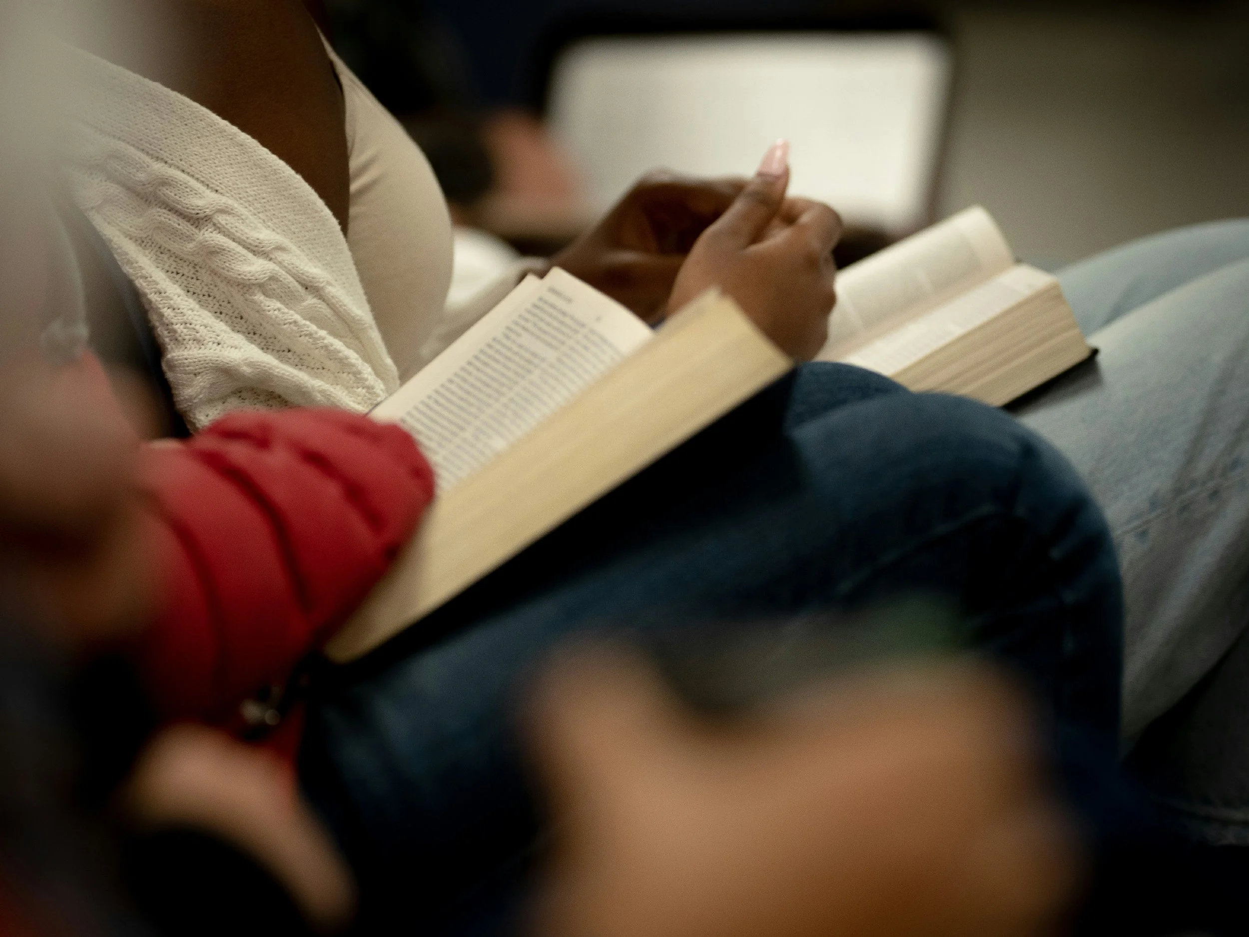 Close-up of a person reading a book, wearing a cream-colored sweater, with other people in the background.