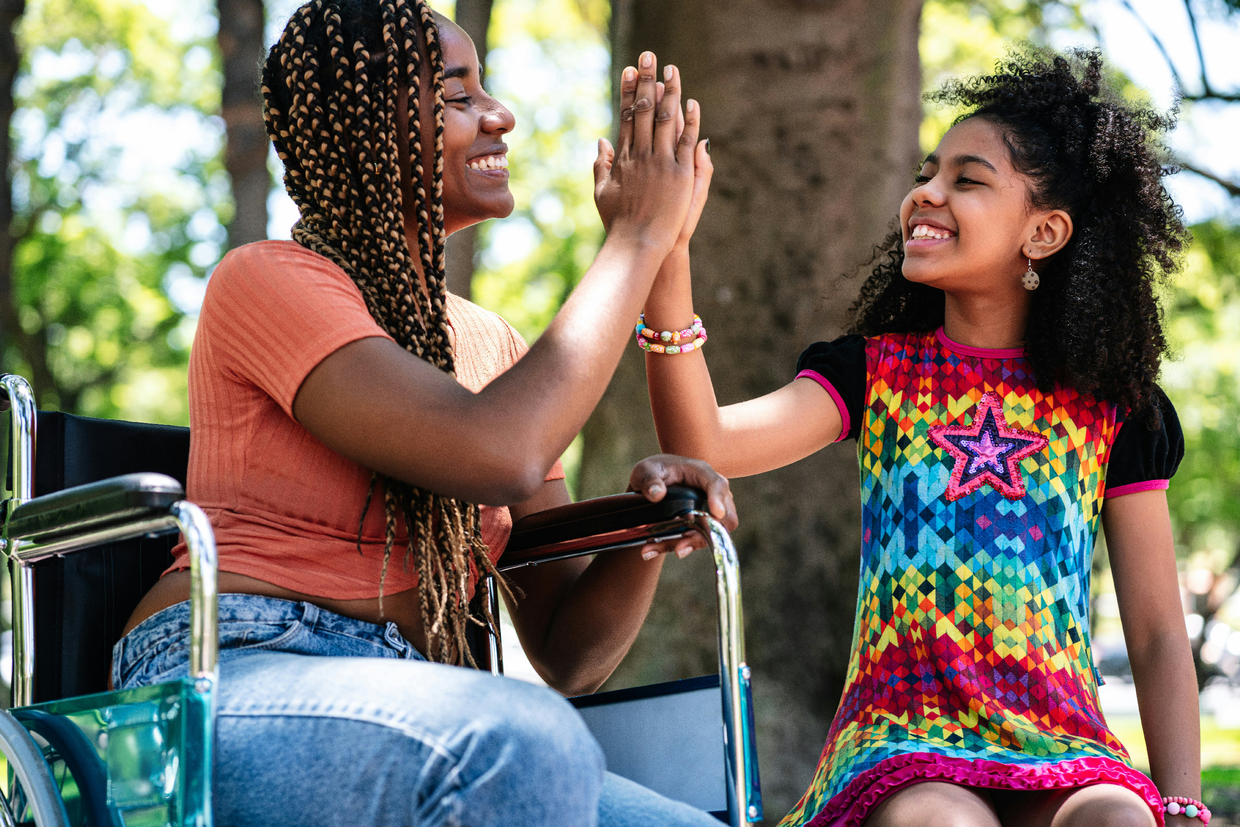 A woman in a wheelchair and a young girl are outdoors, smiling and playing a high-five game in front of a tree.
