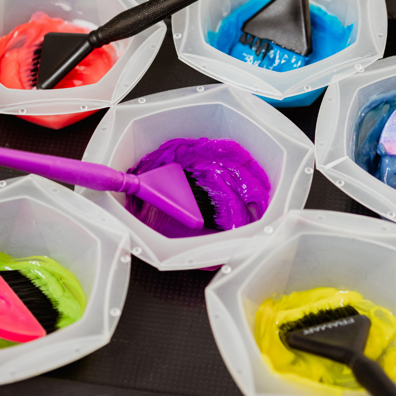 Multiple small containers filled with brightly colored hair dye and small brushes immersed in the dye.