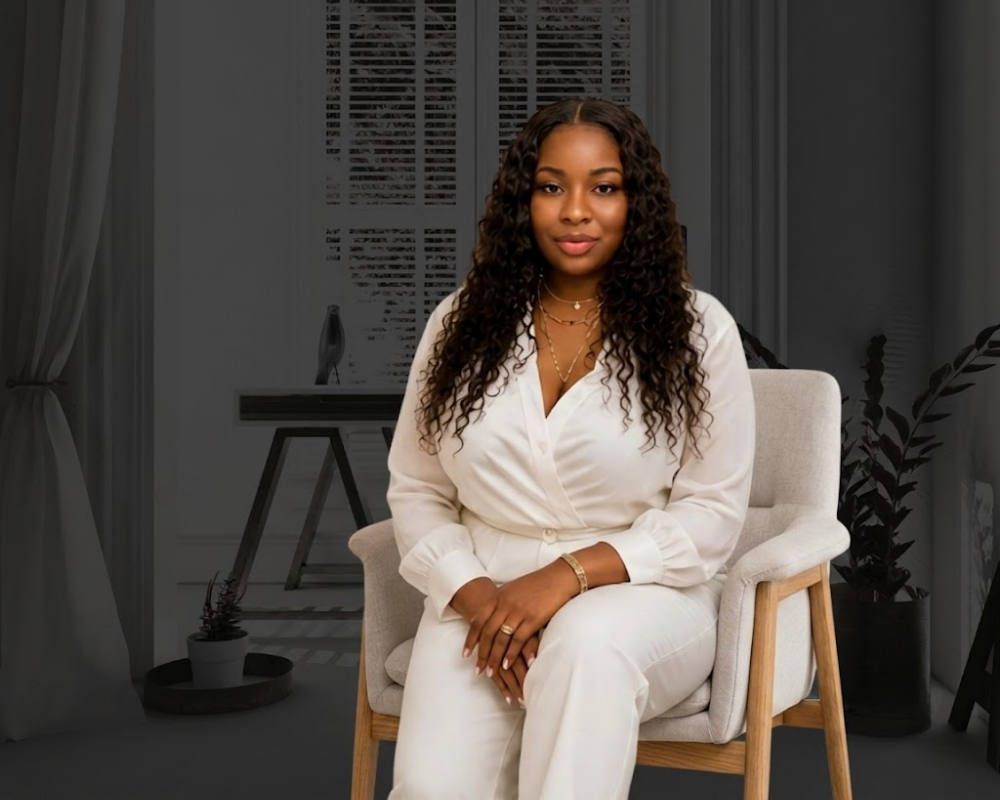 A woman with curly hair and brown skin is sitting on a beige cushioned chair in an indoor setting, wearing a white outfit and gold jewelry, with a calm expression.