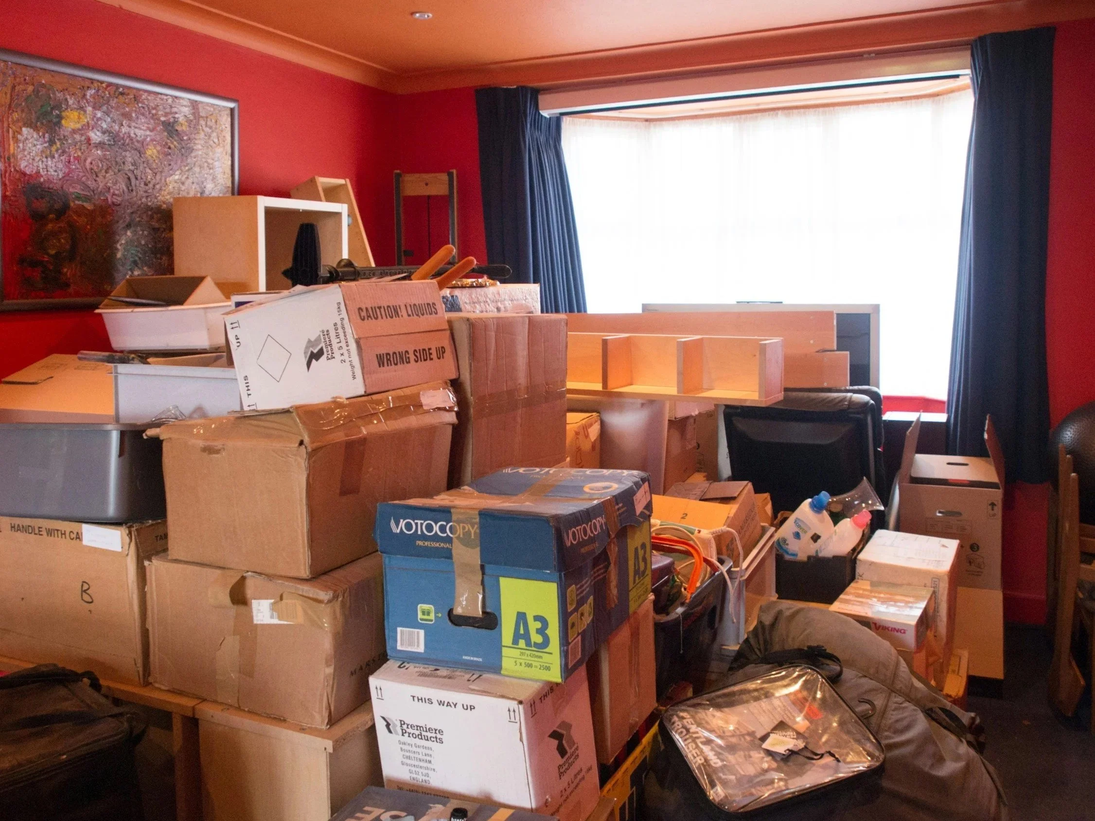 Zimmer mit roten Wänden und Fenster mit blauen Vorhängen. Es gibt gestapelte Pakete und Möbel, die anscheinend aufgebaut werden, einschließlich Kartons, Regalbclocker und Werkzeug. Er scheint sich in einem Raum zu befinden, der für den Umbau oder die Einrichtung vorbereitet wird.