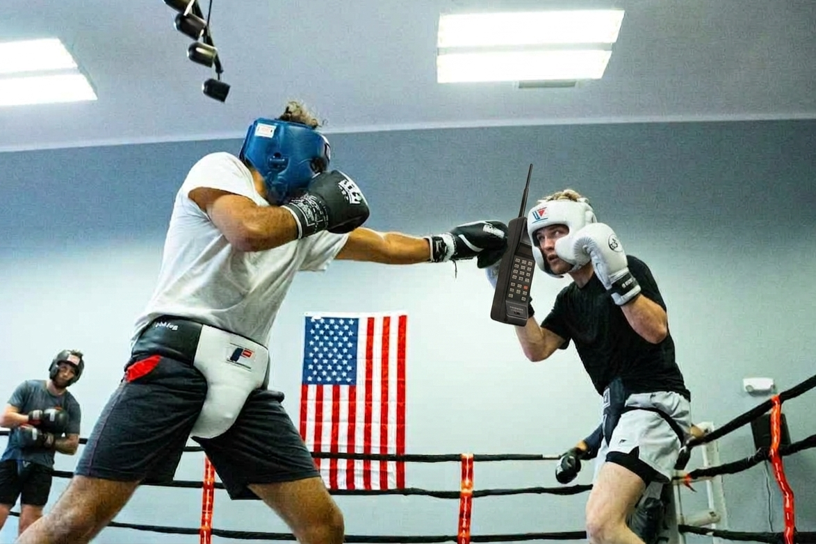 Two men in a boxing ring are sparring, with one wearing a blue helmet and gloves, and the other wearing a white helmet and gloves. The man on the right is holding a cordless phone to his ear. An American flag hangs on the wall in the background.