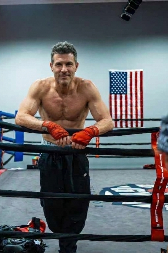 Masters boxer training inside the ring at Central Oregon Boxing in Bend, Oregon.A shirtless man with orange hand wraps leaning on a boxing ring's ropes, smiling, in a gym with an American flag in the background.