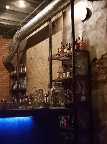 A moody, low-lit photograph of the physical speakeasy bar interior, showing glowing warm light on rows of glass liquor bottles against a rustic brick wall.