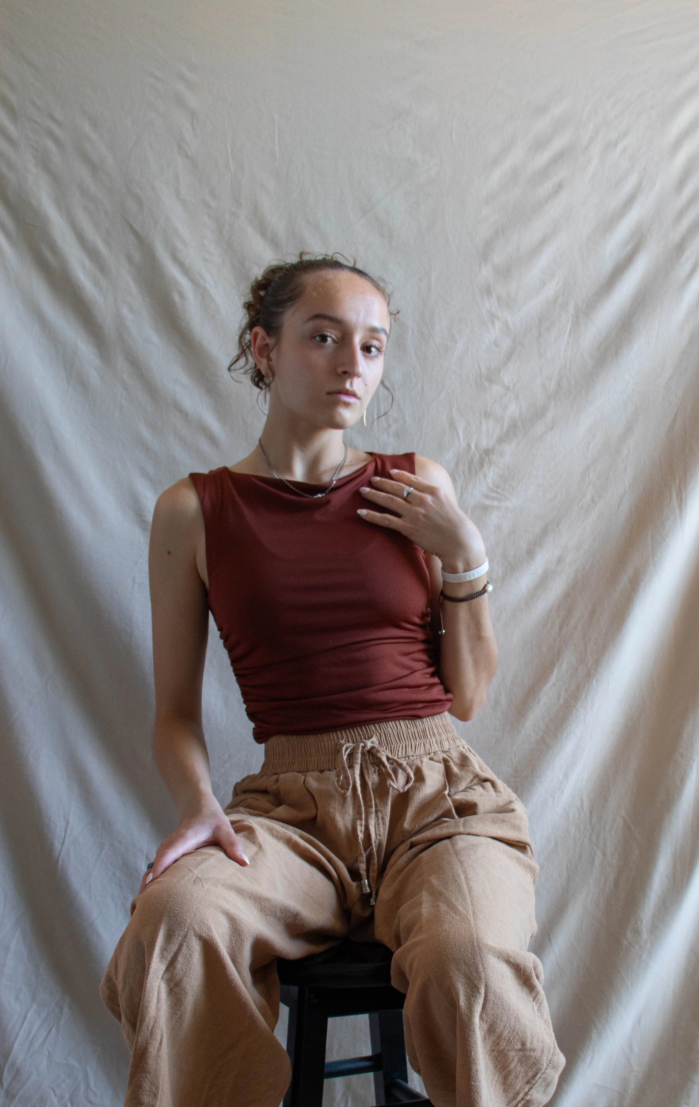 A young woman with curly hair, wearing a sleeveless rust-colored top and beige drawstring pants, sitting on a black chair against a beige fabric backdrop, looking directly at the camera with a neutral expression.