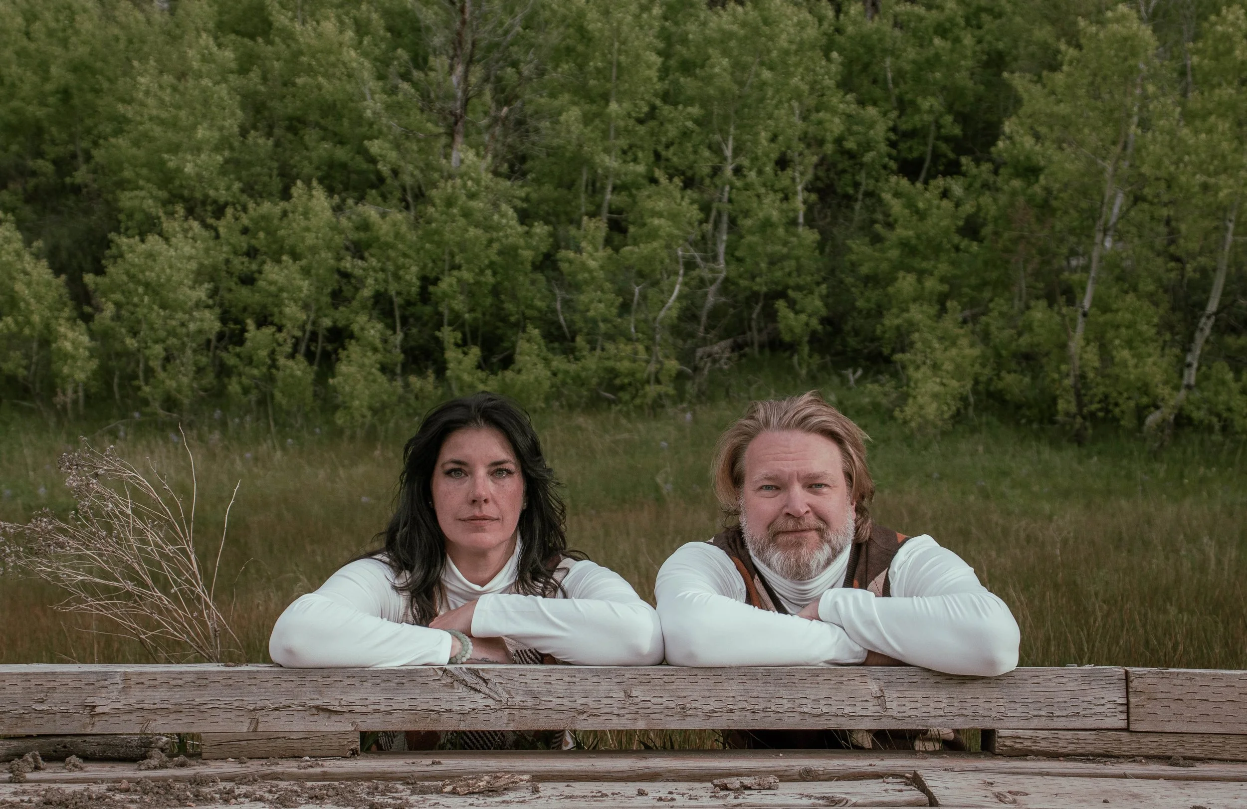 Two people, a woman with dark hair and a man with light hair and a beard, are sitting behind a weathered wooden fence outdoors, with a background of green trees and grass.