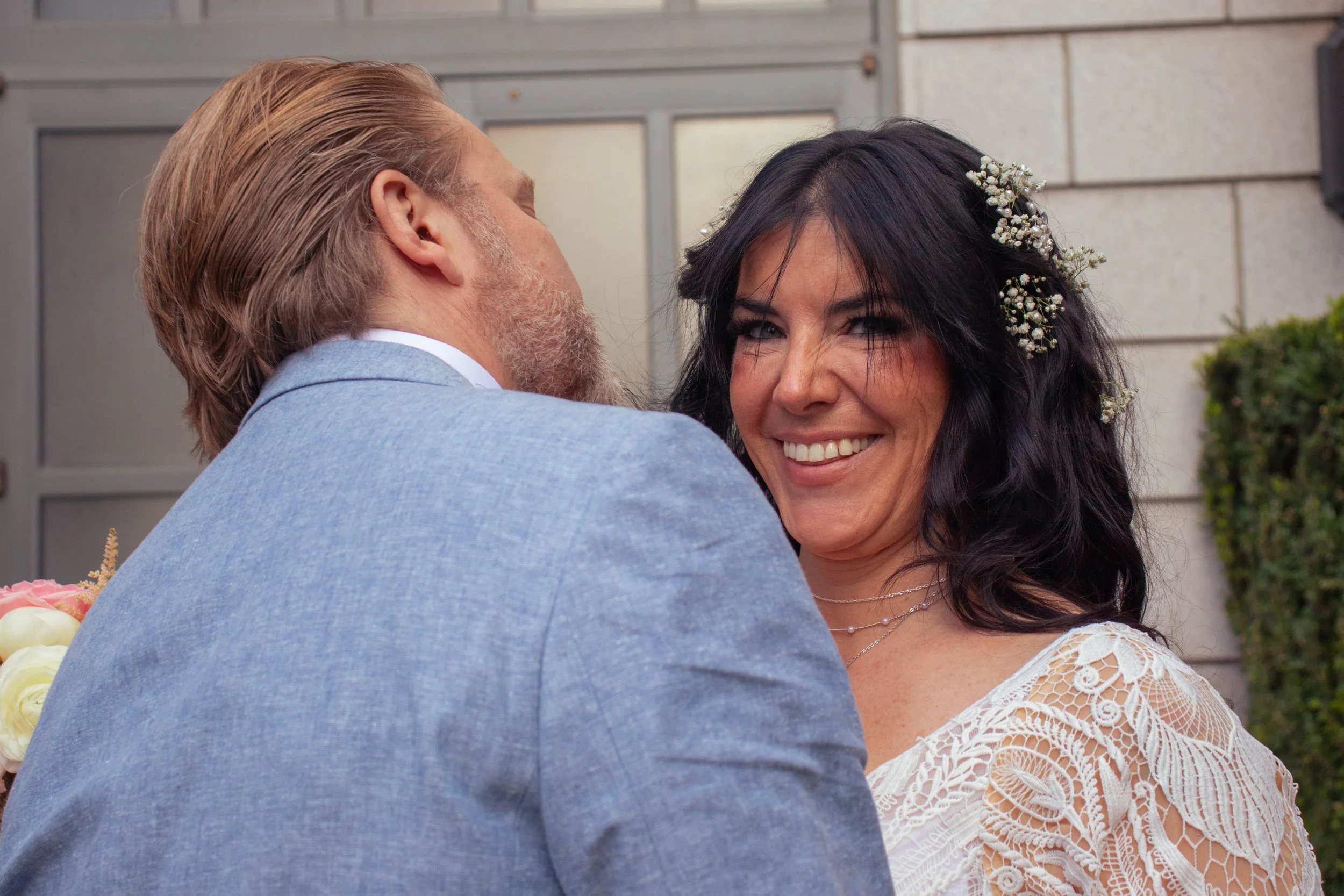 A man and woman in a close embrace, with the woman smiling, wedding attire, with hair decorated with small white flowers, standing outdoors near a building.