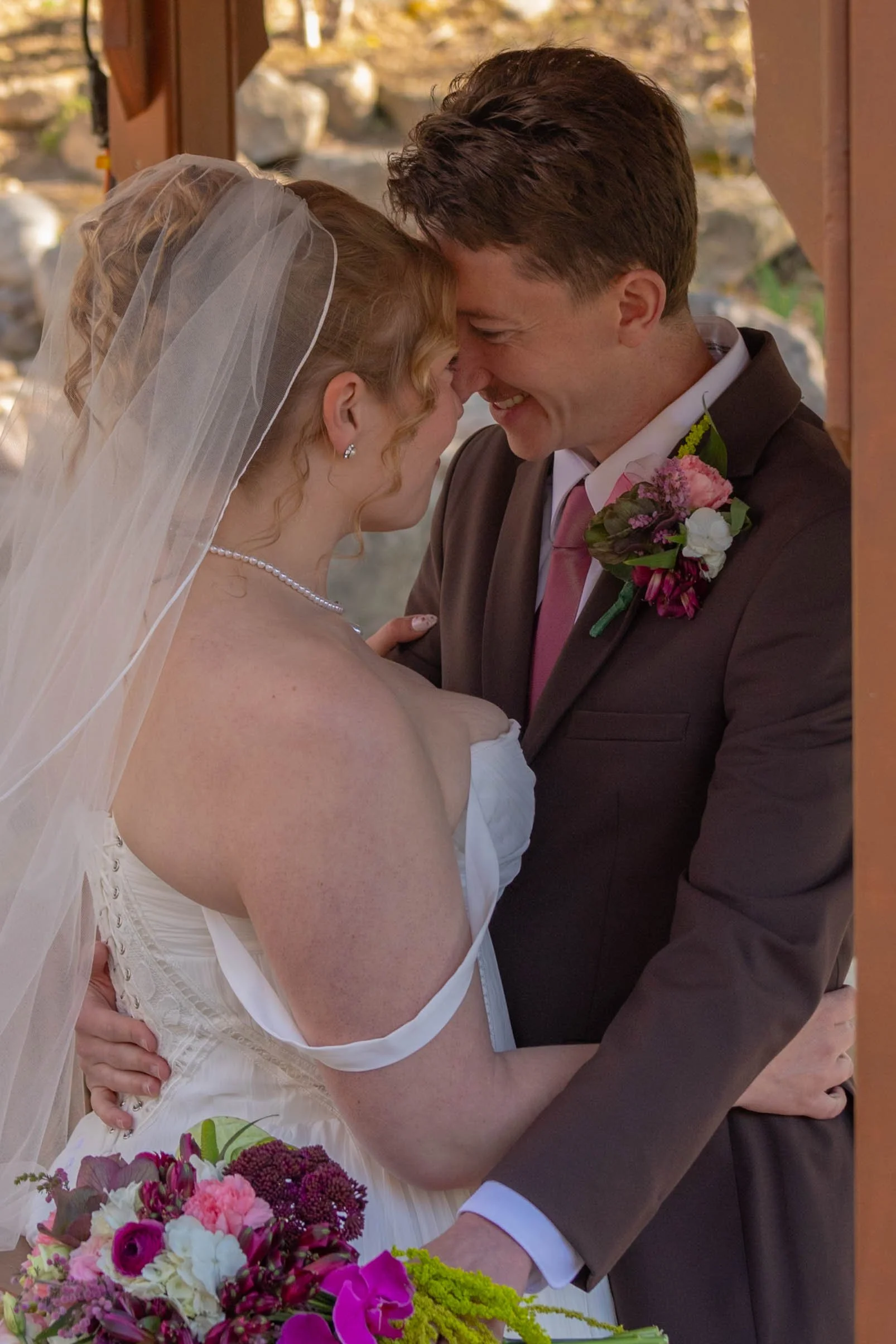 A newlywed couple is sharing a close, joyful moment during their wedding, with their foreheads touching and eyes closed, surrounded by colorful flowers.
