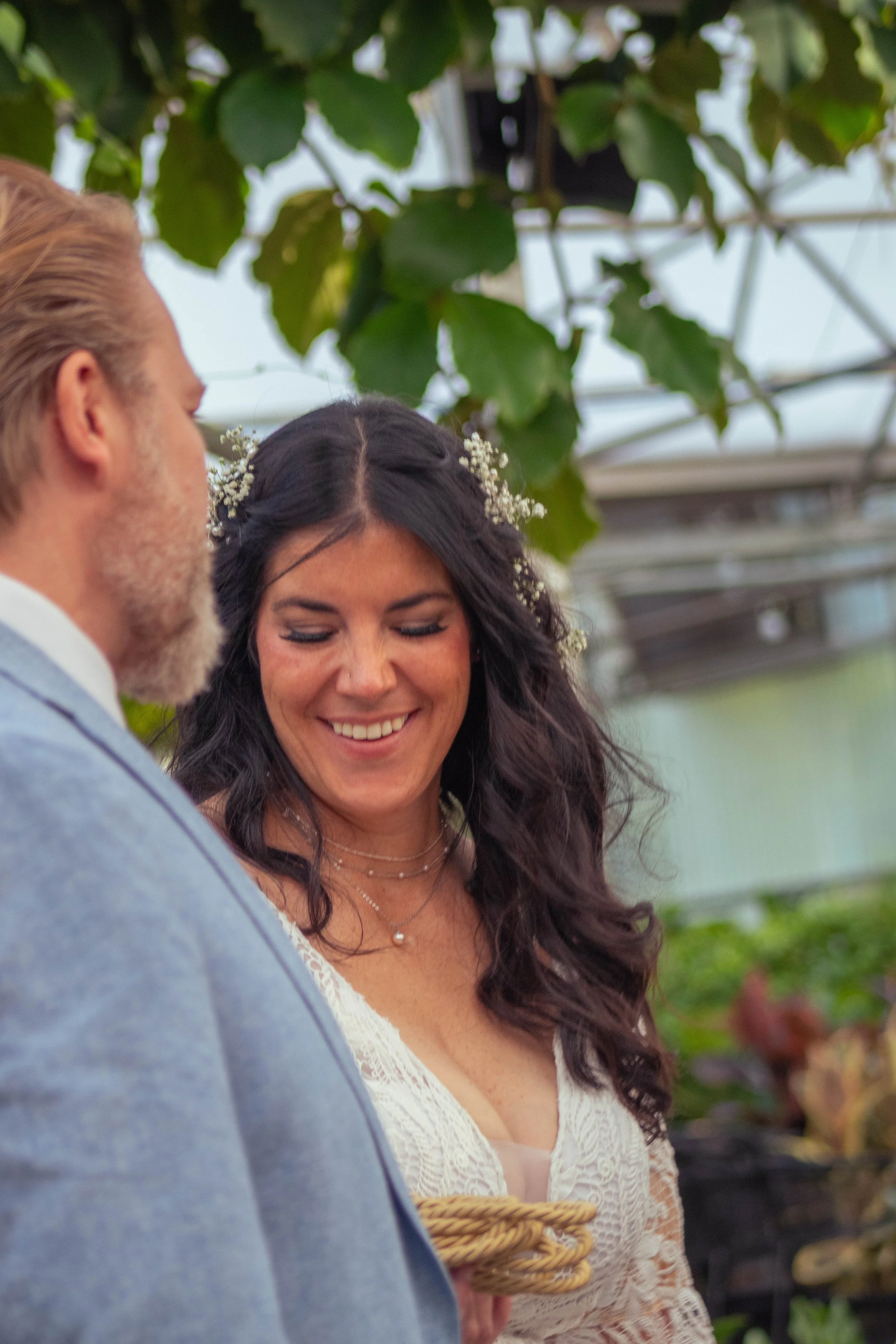 Close-up of a smiling woman with long dark hair decorated with small white flowers, standing outdoors near greenery, wearing a white lace dress and layered necklaces.