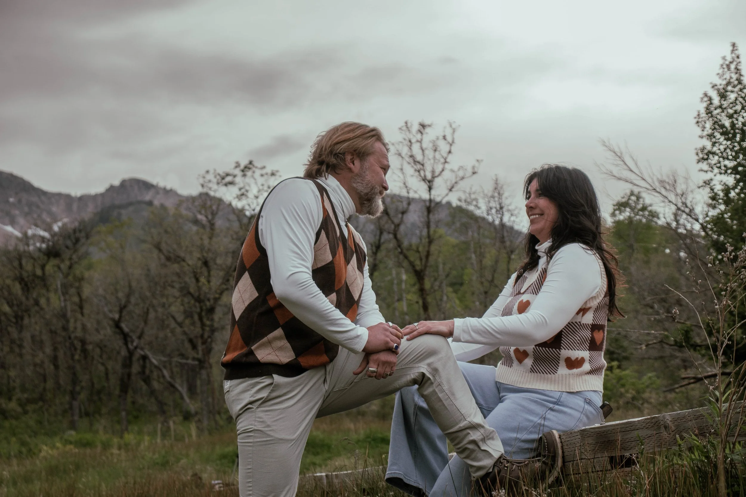 A couple dates outdoors, with the man kneeling and holding the woman's hands, as she smiles. They are surrounded by trees, grass, and mountains in the background.